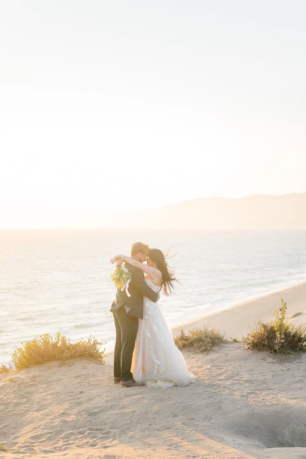 Bride and groom embrace on a sandy bluff at Point Dume during golden hour, bathed in warm sunlight with the Pacific Ocean and distant hills softly glowing in the background.