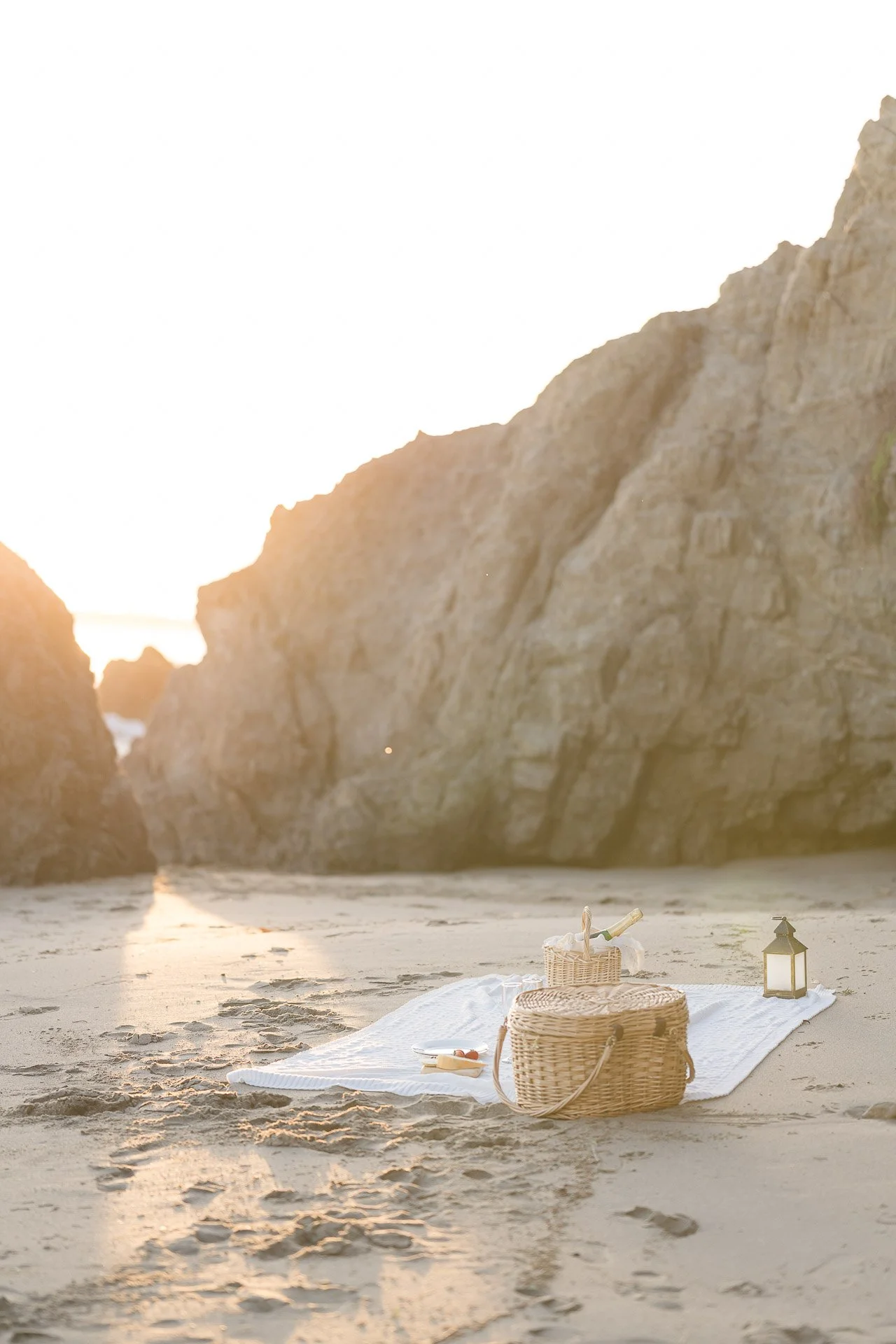 Summer Engagement Photos in Malibu featuring a romantic beach picnic with wicker baskets, champagne, and a lantern surrounded by dramatic coastal cliffs at sunset.