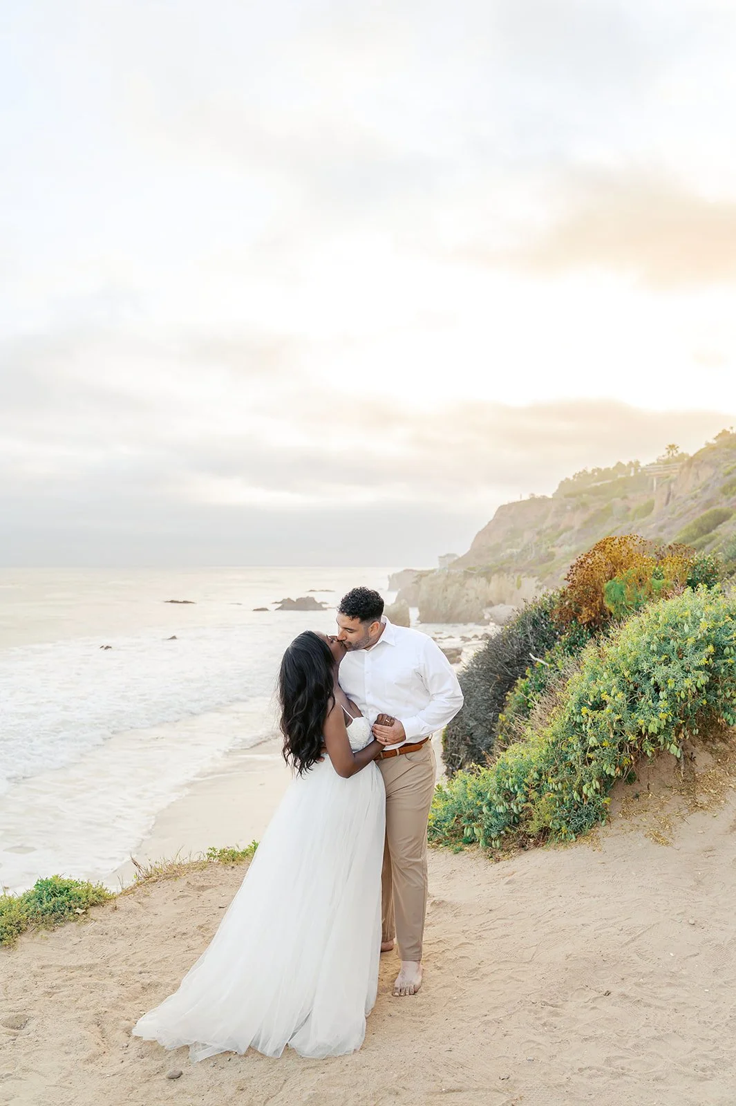Newlyweds share a kiss on a sandy cliffside above a peaceful beach, with lush greenery and coastal bluffs behind them at sunset. This romantic oceanfront location highlights why it’s considered one of the Best Places to Elope 2026.