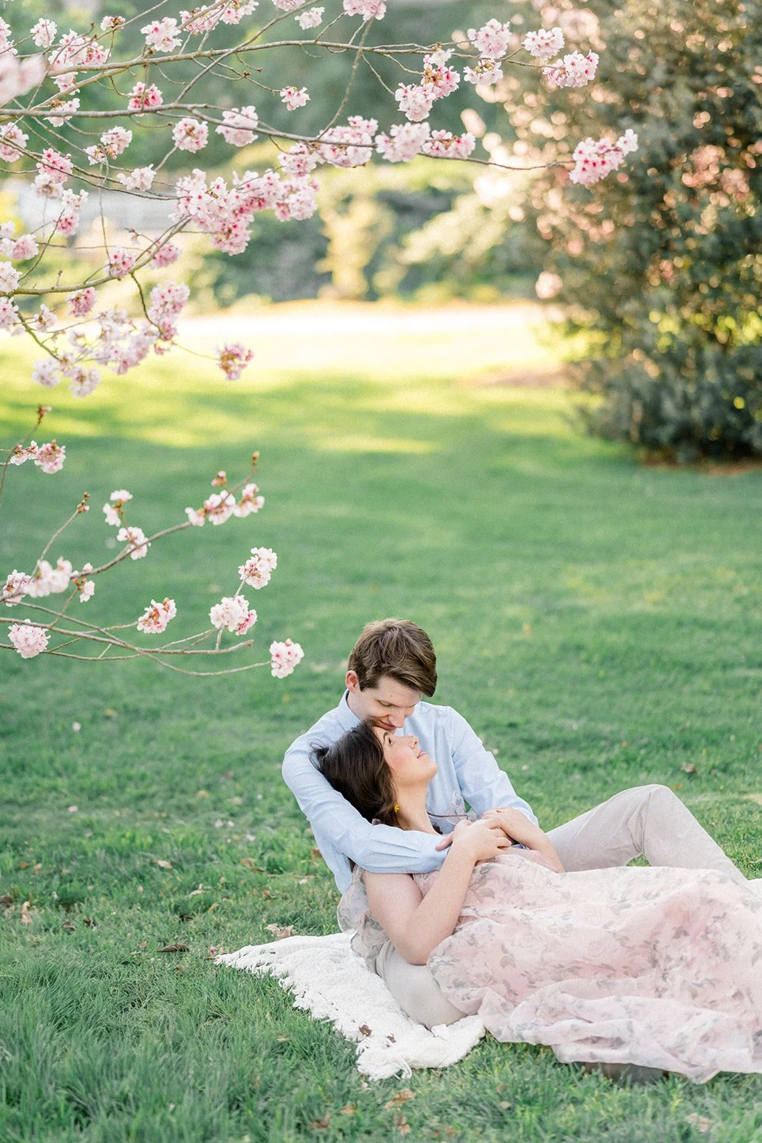 Cherry Blossom Photoshoot ideas featuring a couple lying on a blanket beneath blooming cherry blossom branches, cuddled close as they smile at each other in soft, golden spring light