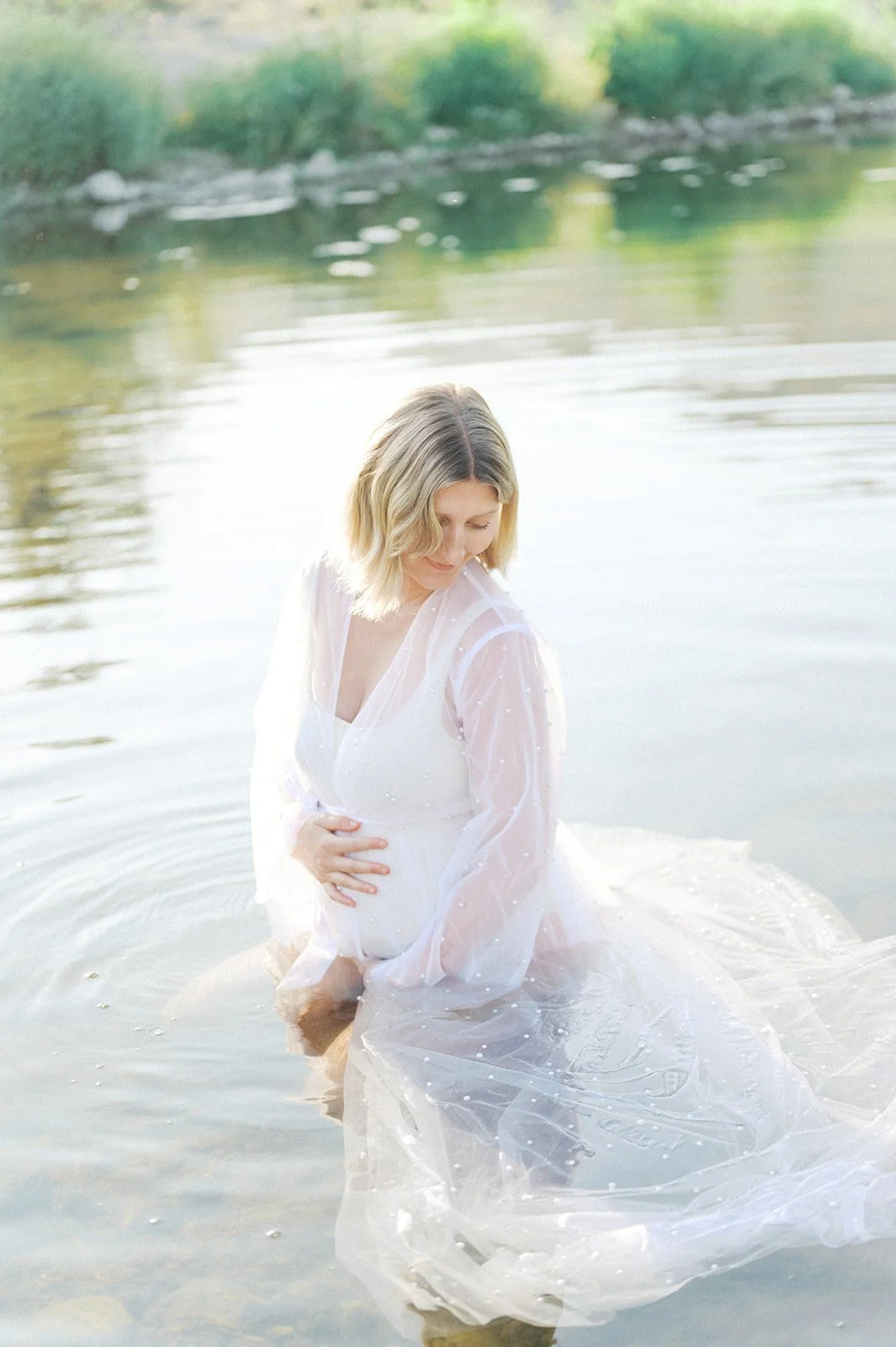 Summer Maternity Photos of an expecting mother sitting in a calm river wearing a flowing sheer white gown during soft golden evening light.