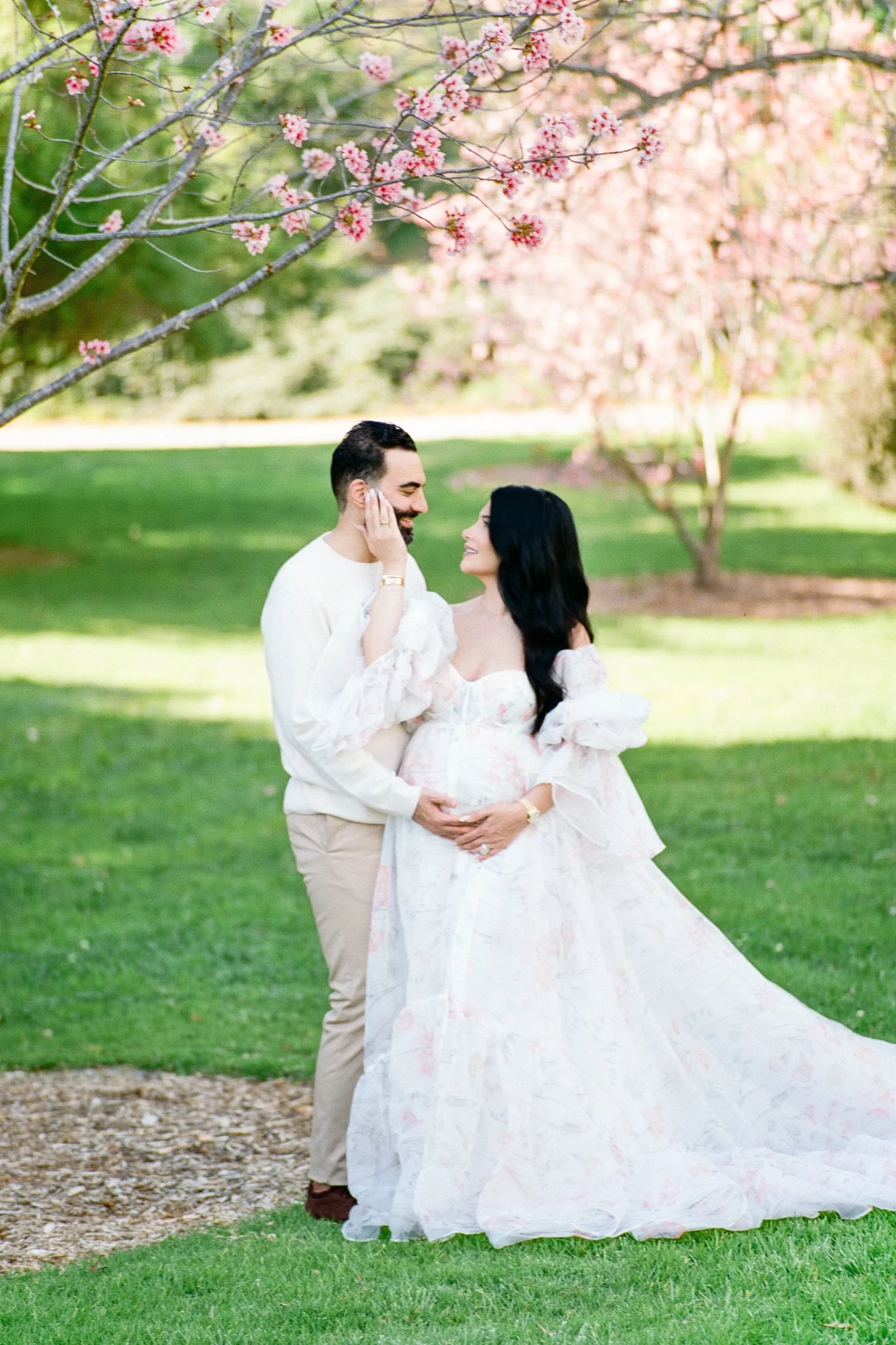 Cherry blossom Maternity photos of an expecting couple standing beneath blooming pink trees, smiling at each other while gently holding the baby bump in soft, natural spring light.