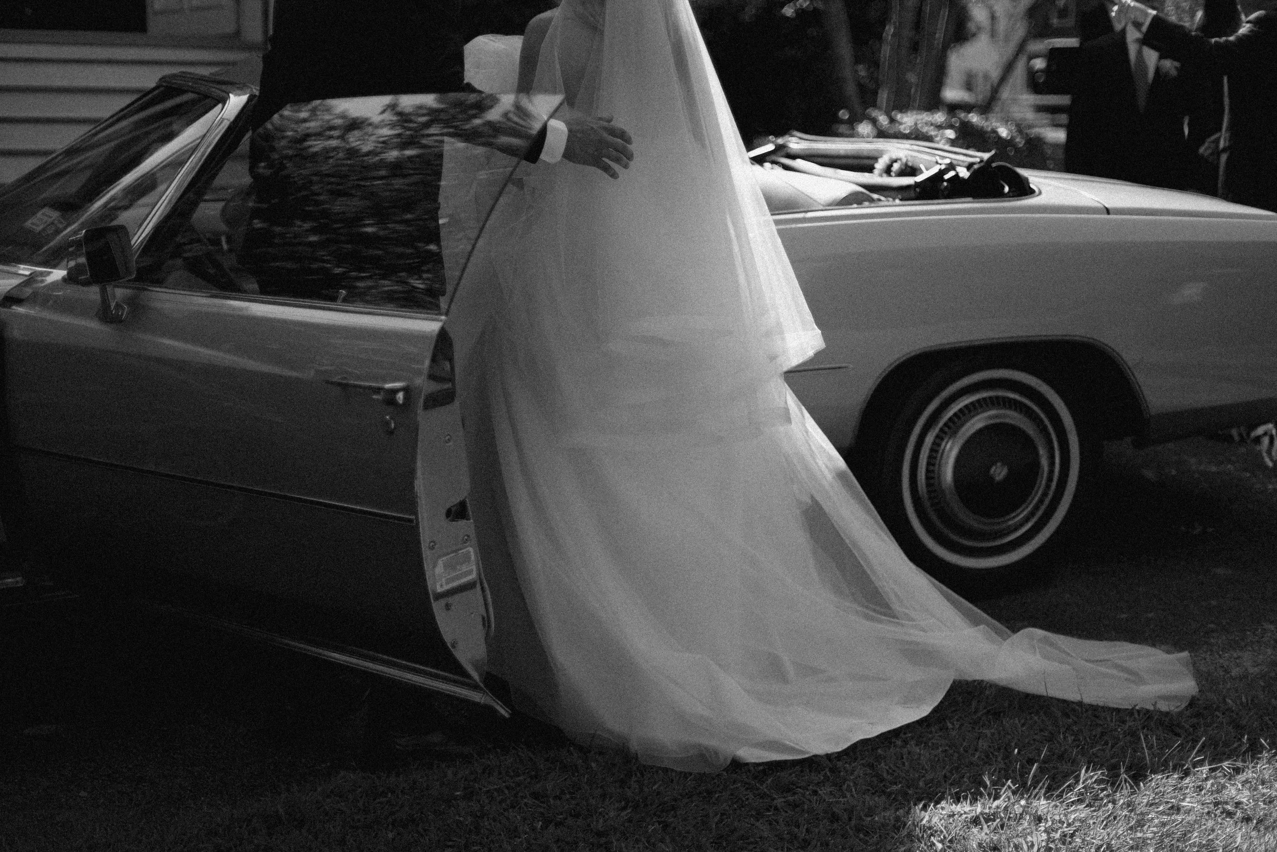 A bride in a wedding gown standing next to a vintage convertible car, with the car door open, on a grassy area.