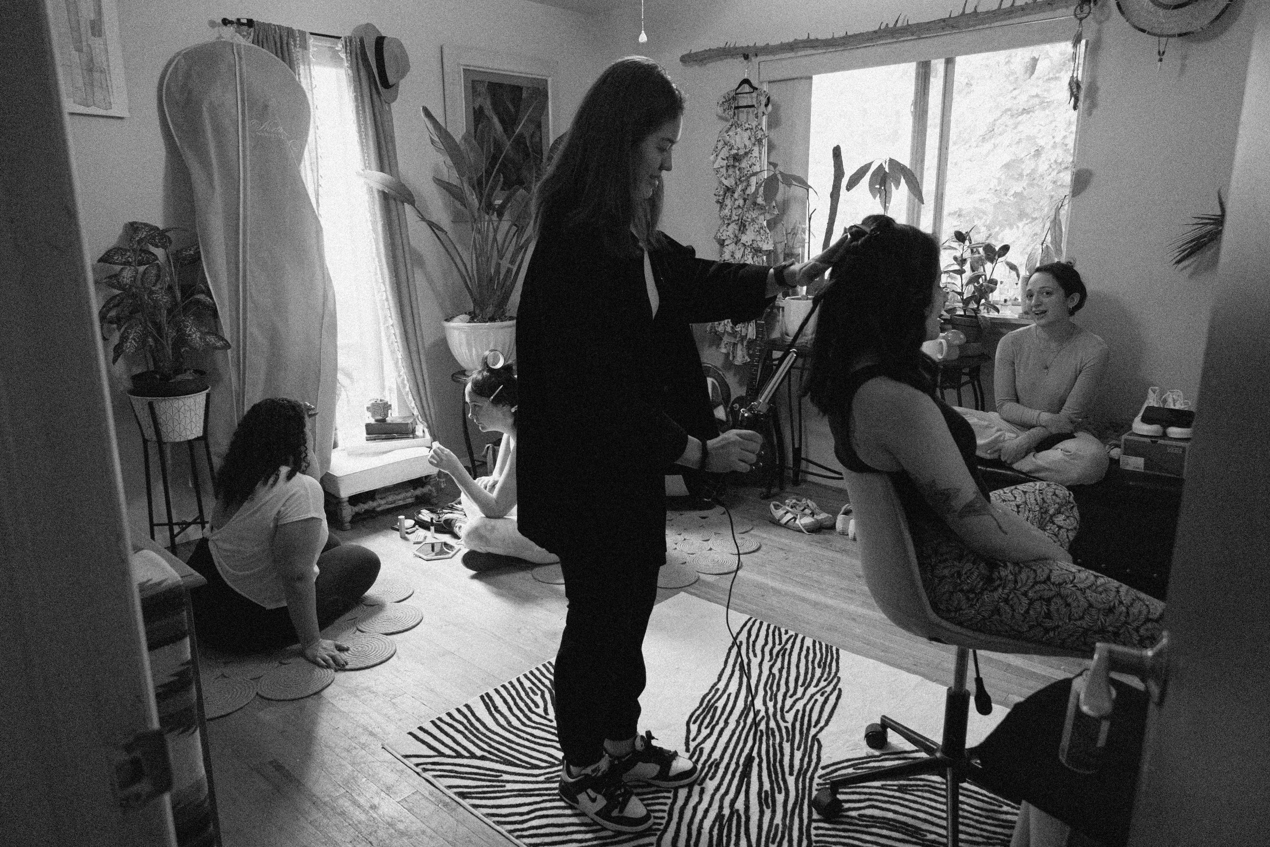 A bride getting her hair styled by a hairstylist in a cozy room filled with plants, with two other women sitting on the floor and on a chair in the background. Getting ready bridal portraits. 