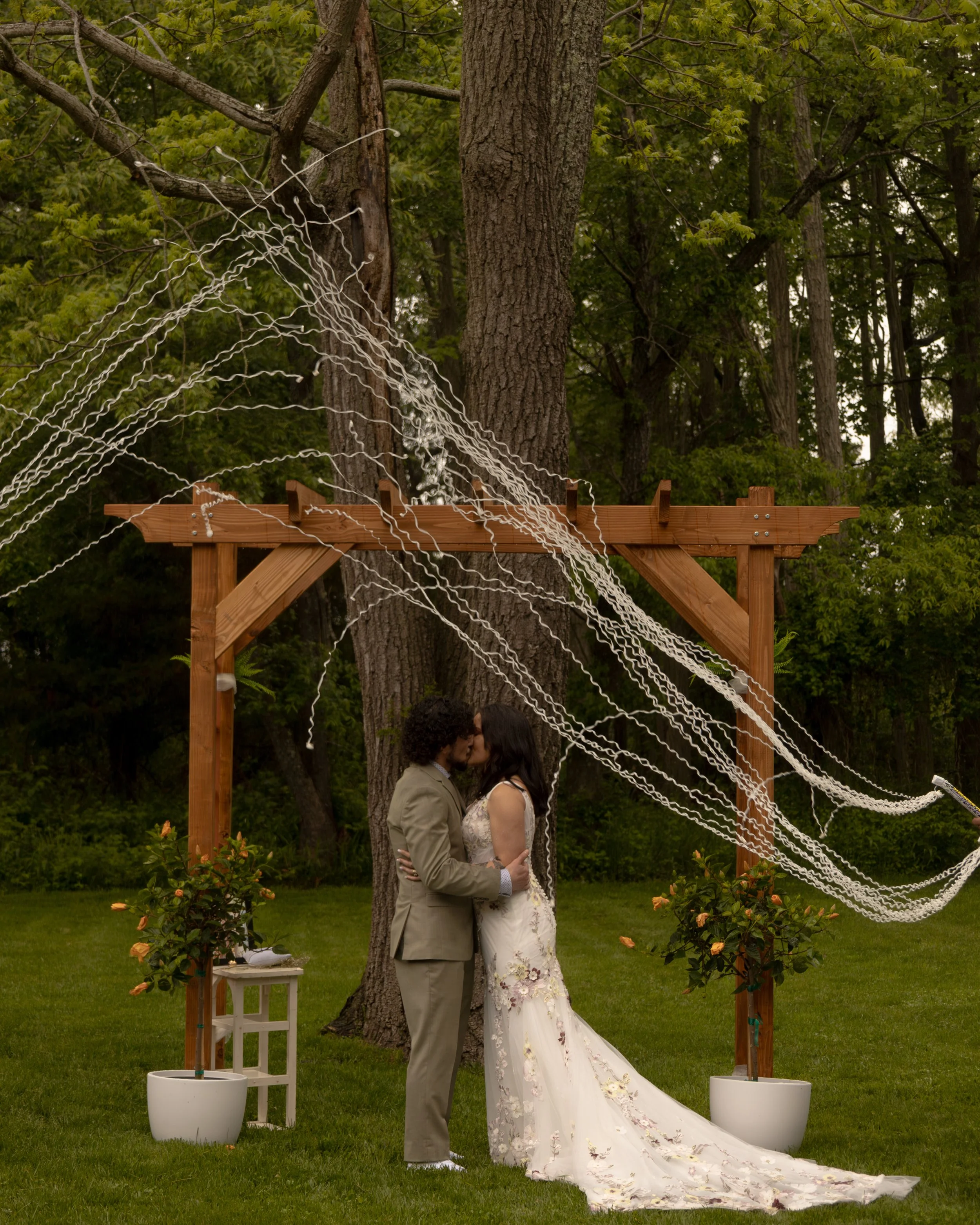 A couple kissing outdoors during a wedding ceremony under a wooden arch decorated with white ribbons, with trees and grass in the background. Romantic north fork wedding photographer. 