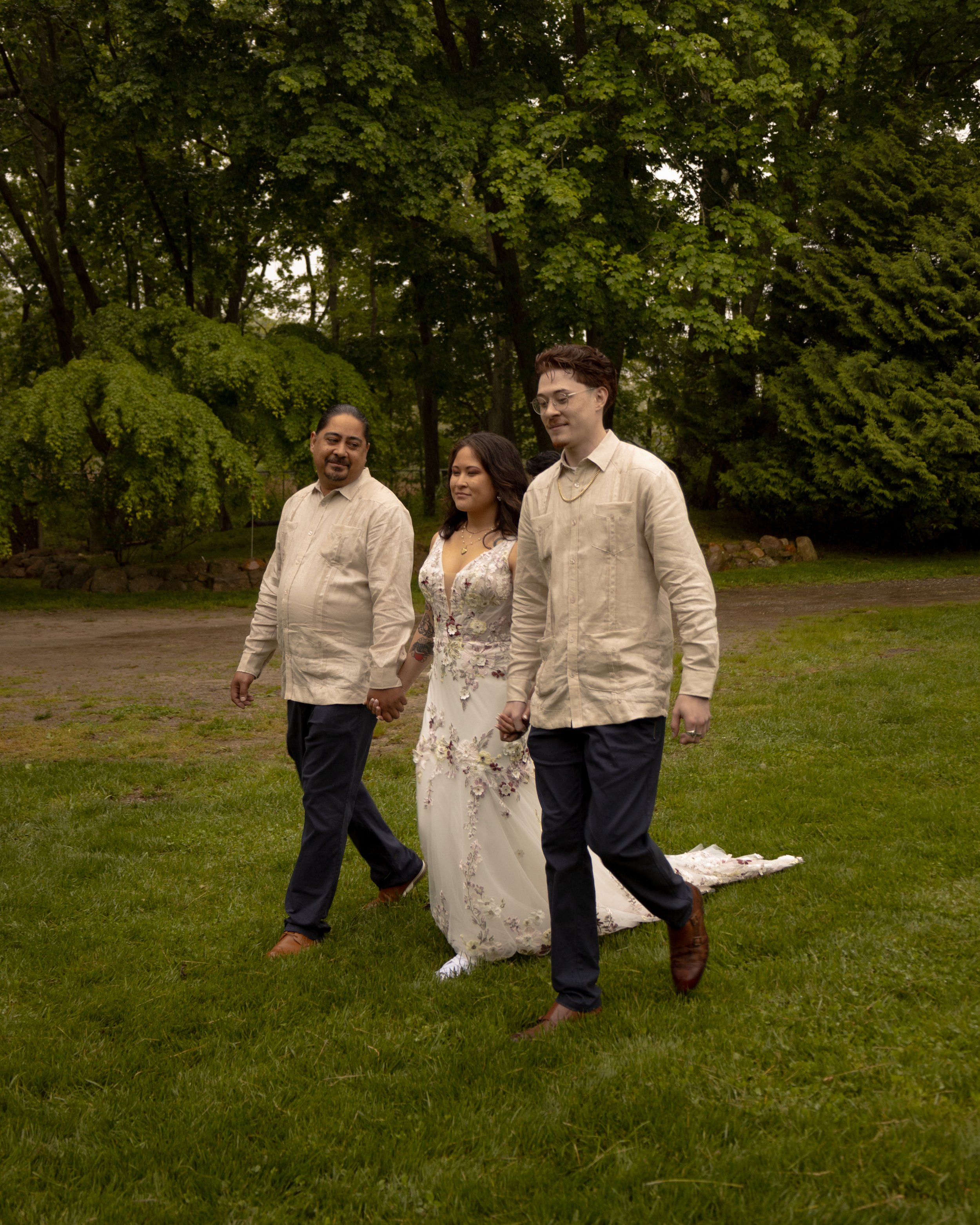 Three people holding hands in a park during a wedding ceremony, with trees and grass in the background. Greenport Wedding Photographer. Mattituck Wedding Photographer. Southold Wedding Photographer. 