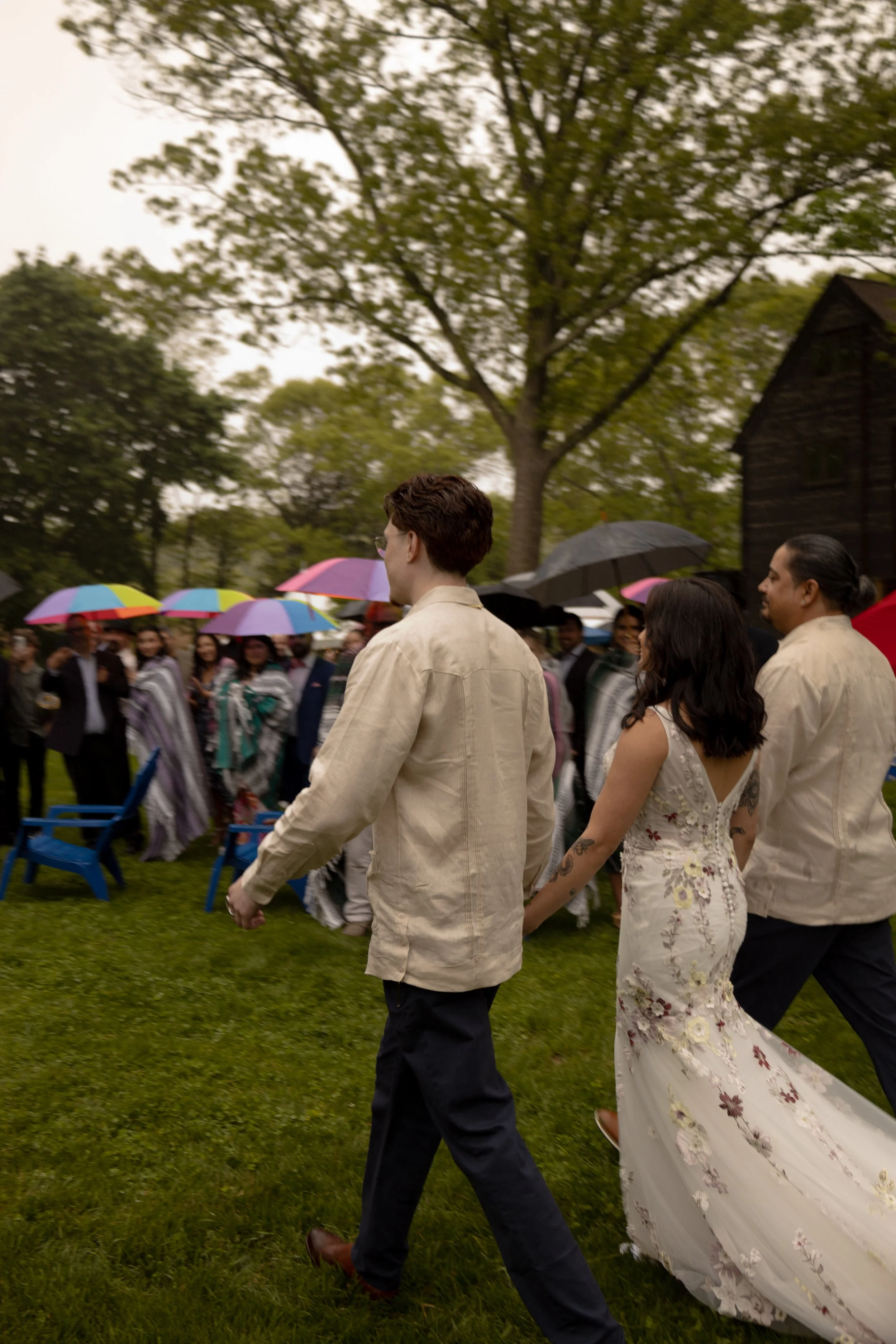 People attending an outdoor wedding, walking hand in hand on a grassy lawn, with some holding colorful umbrellas, trees and a wooden building in the background. Long Island Wedding Photographer. 