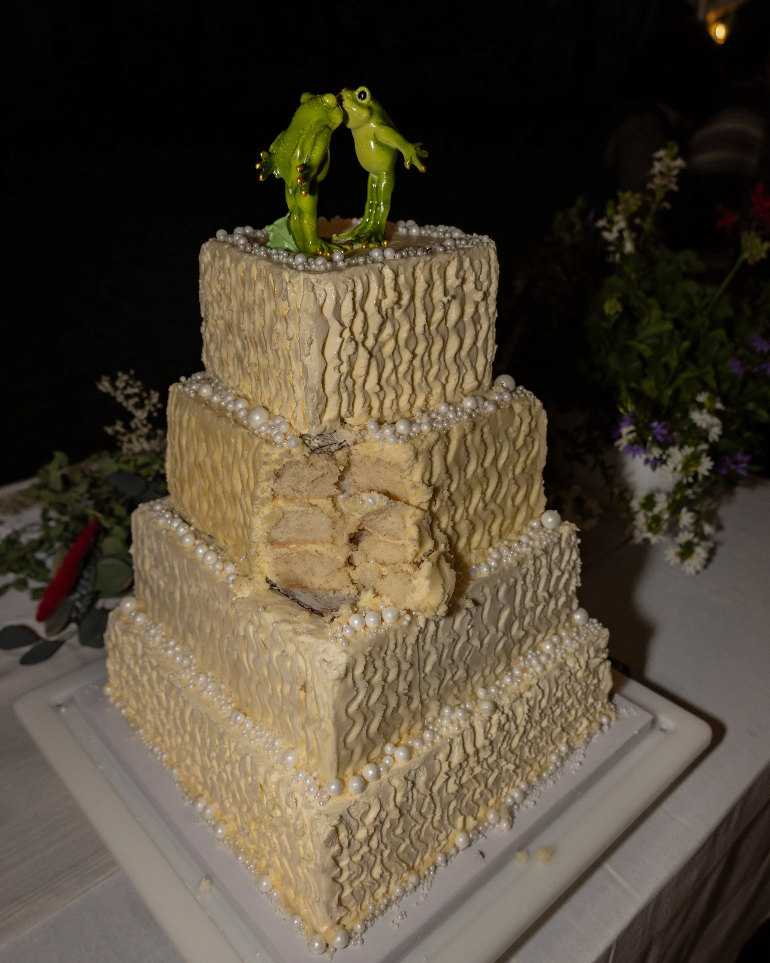 A multi-tiered wedding cake with beige textured fondant, decorated with white edible pearls and topped with two green frog figurines kissing, set against a dark background with some flowers. Editorial north fork wedding photographer. Editorial. 