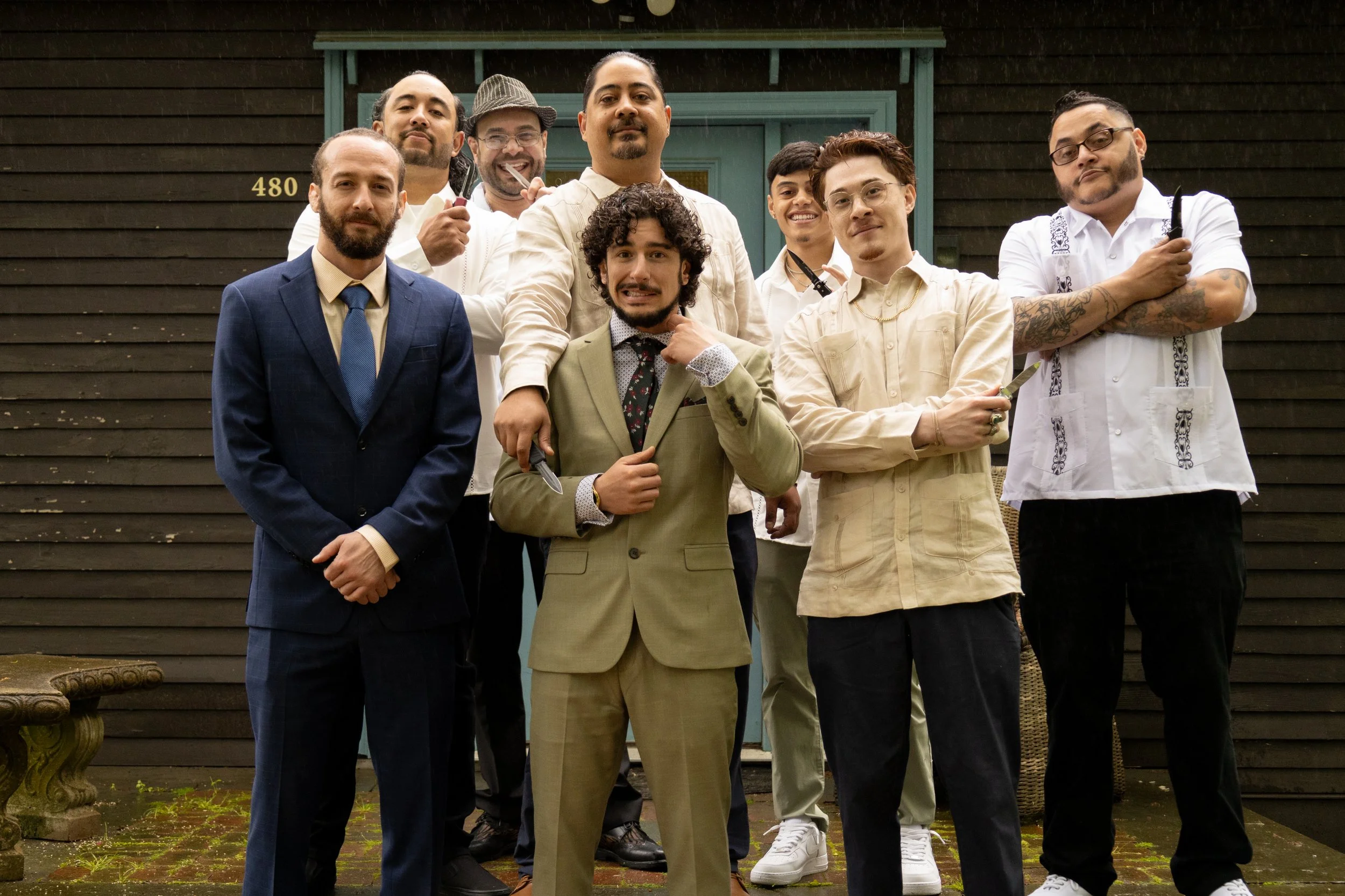 A group of nine men pose in front of a house, some holding knives and other tools, with one man in a suit, and some with tattoos and shaved heads, standing on a mossy brick patio. Groomsmen at a North Fork Wedding. Long Island Wedding Photographer. 