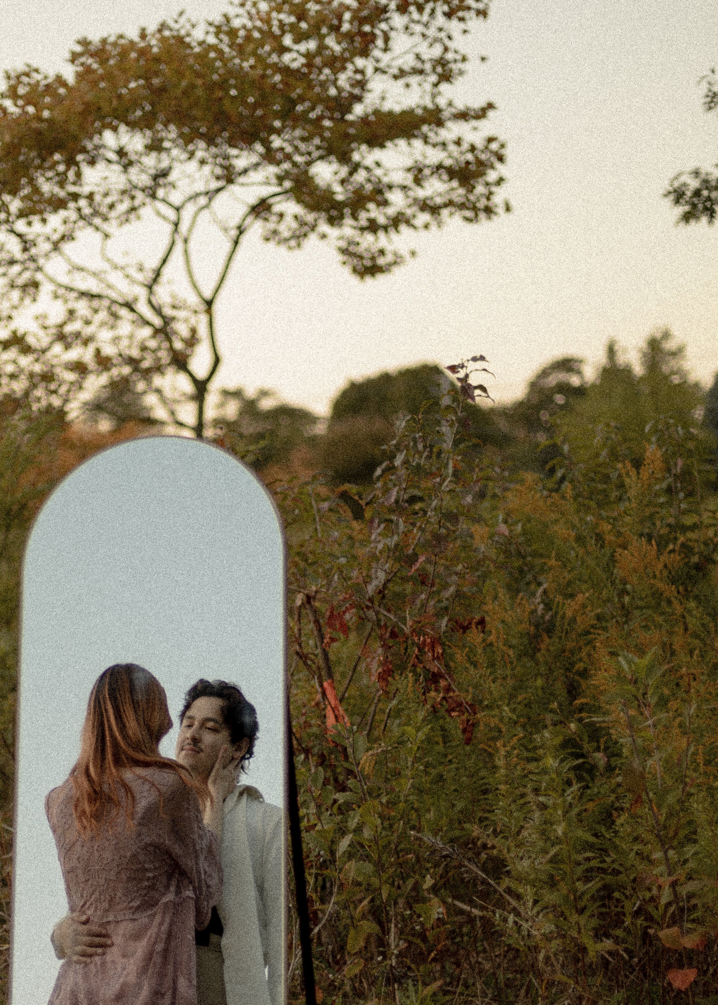 A photograph of a woman and a man embracing outdoors during sunset, with a tall mirror reflecting the couple and the sky with trees in the background.