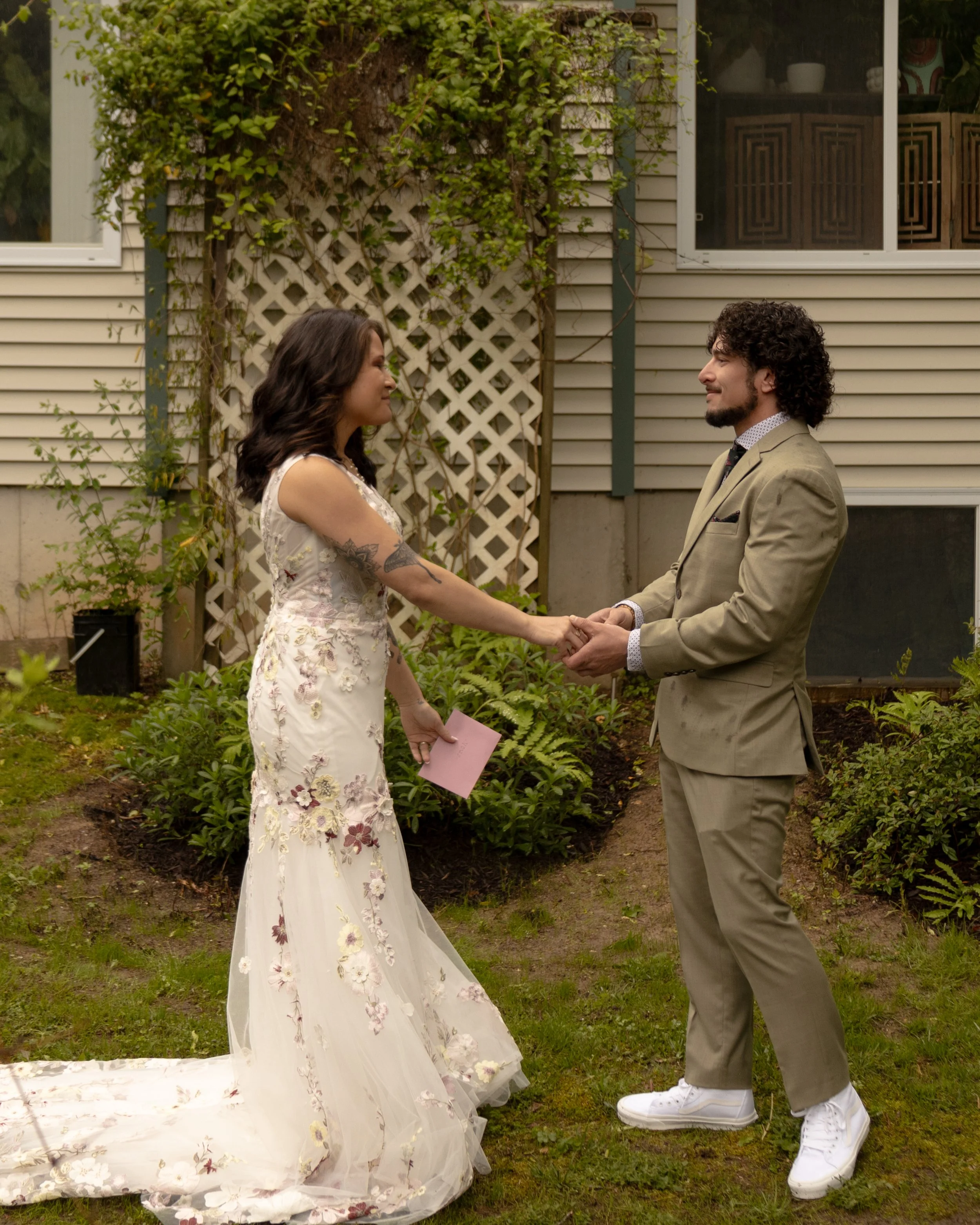 A woman in a white floral wedding dress and a man in a beige suit holding hands and facing each other outdoors, surrounded by green plants and garden elements. Highly recommended north fork wedding photographer. Hamptons wedding photographer. 