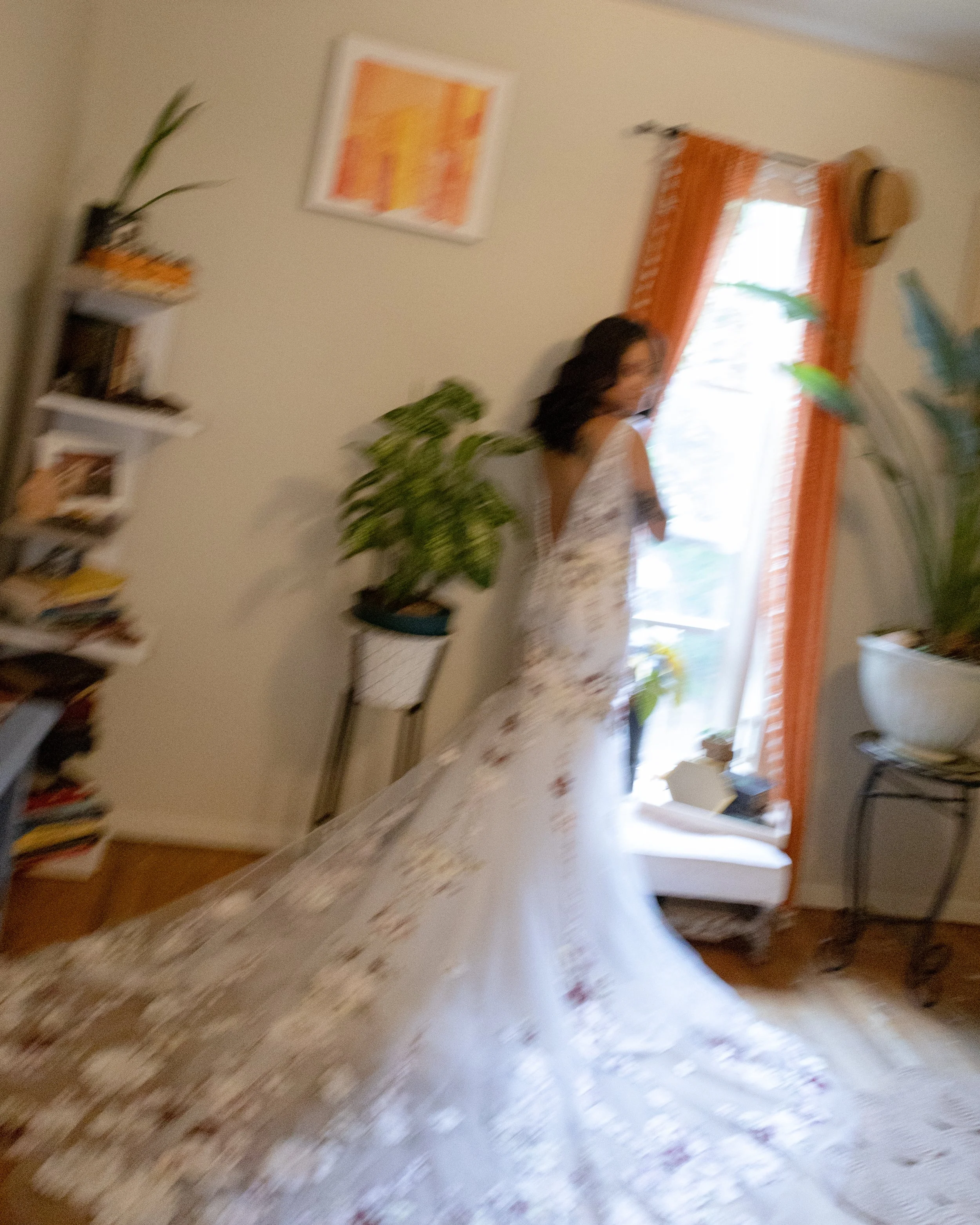 A blurry photo of a woman in a floral dress standing in a living room with orange curtains, a white table, a tall shelf, and several plants. Boho North fork wedding photographer. 