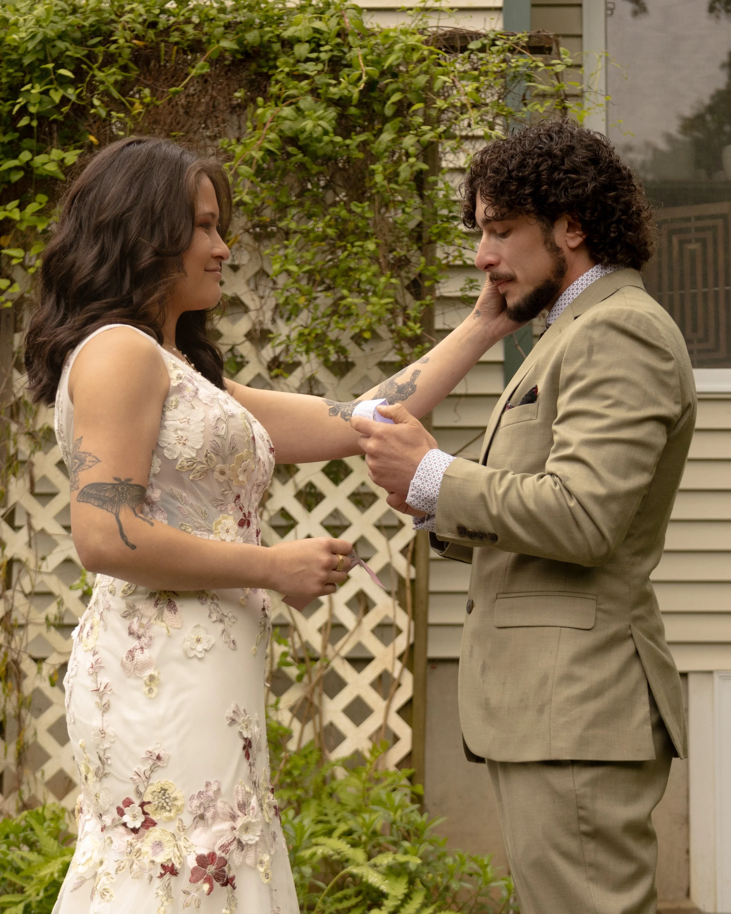 A woman in a floral dress is holding the hand of a man in a tan suit, seemingly during an outdoor wedding ceremony in a garden setting.
