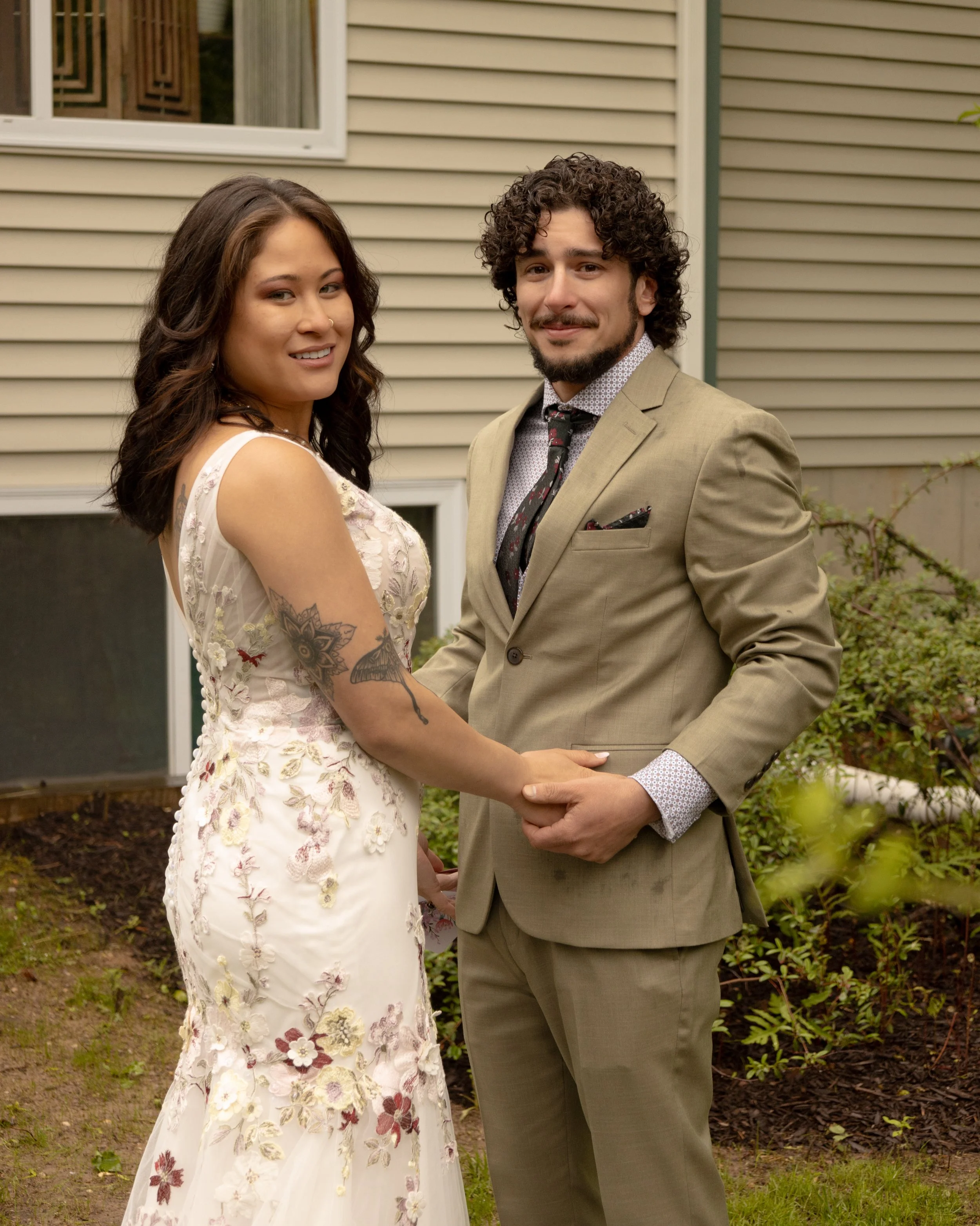 A couple stands outdoors holding hands and smiling at the camera, with a house in the background. East End Wedding Photographer. North Fork Wedding Photographer. Long Island Wedding Photographer. 