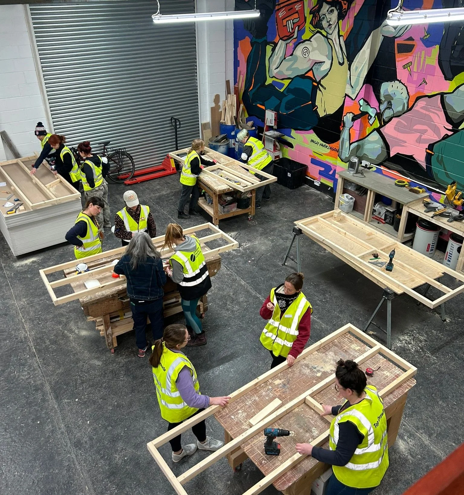 A group of people wearing safety vests are working on wooden frames in a workshop.