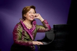 Patricia Goodson smiling at a black piano against a purple background, wearing a floral top with purple accents.
