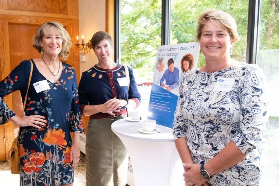 Three women standing beside a small table with coffee cups, at an indoor event, with a large window showing trees outside.