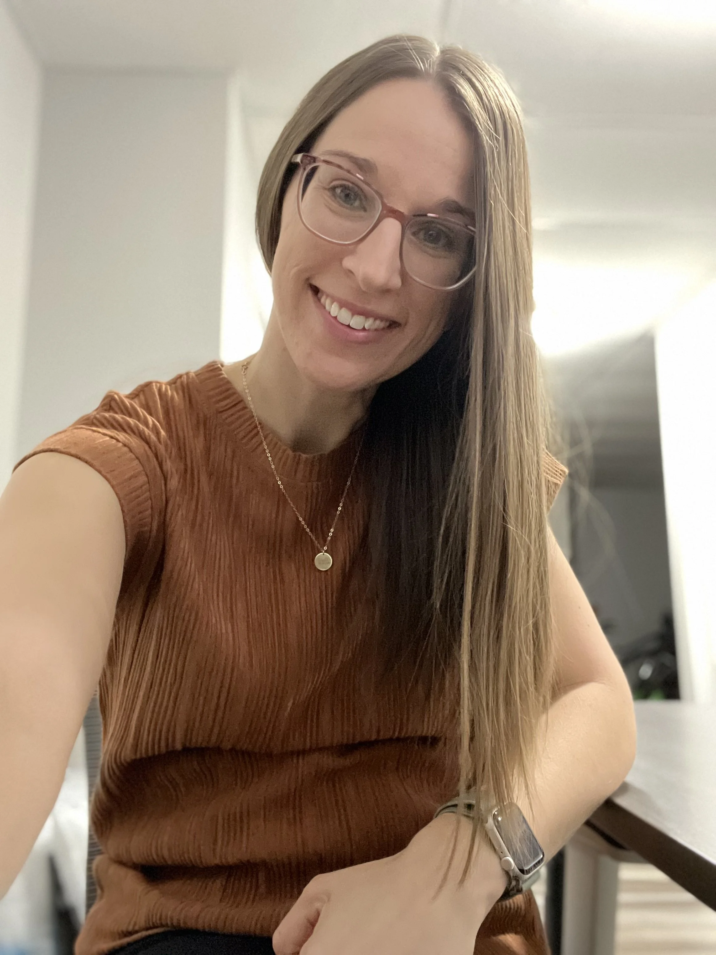 A woman with long brown hair, glasses, and a warm smile sitting at a table inside a home.
