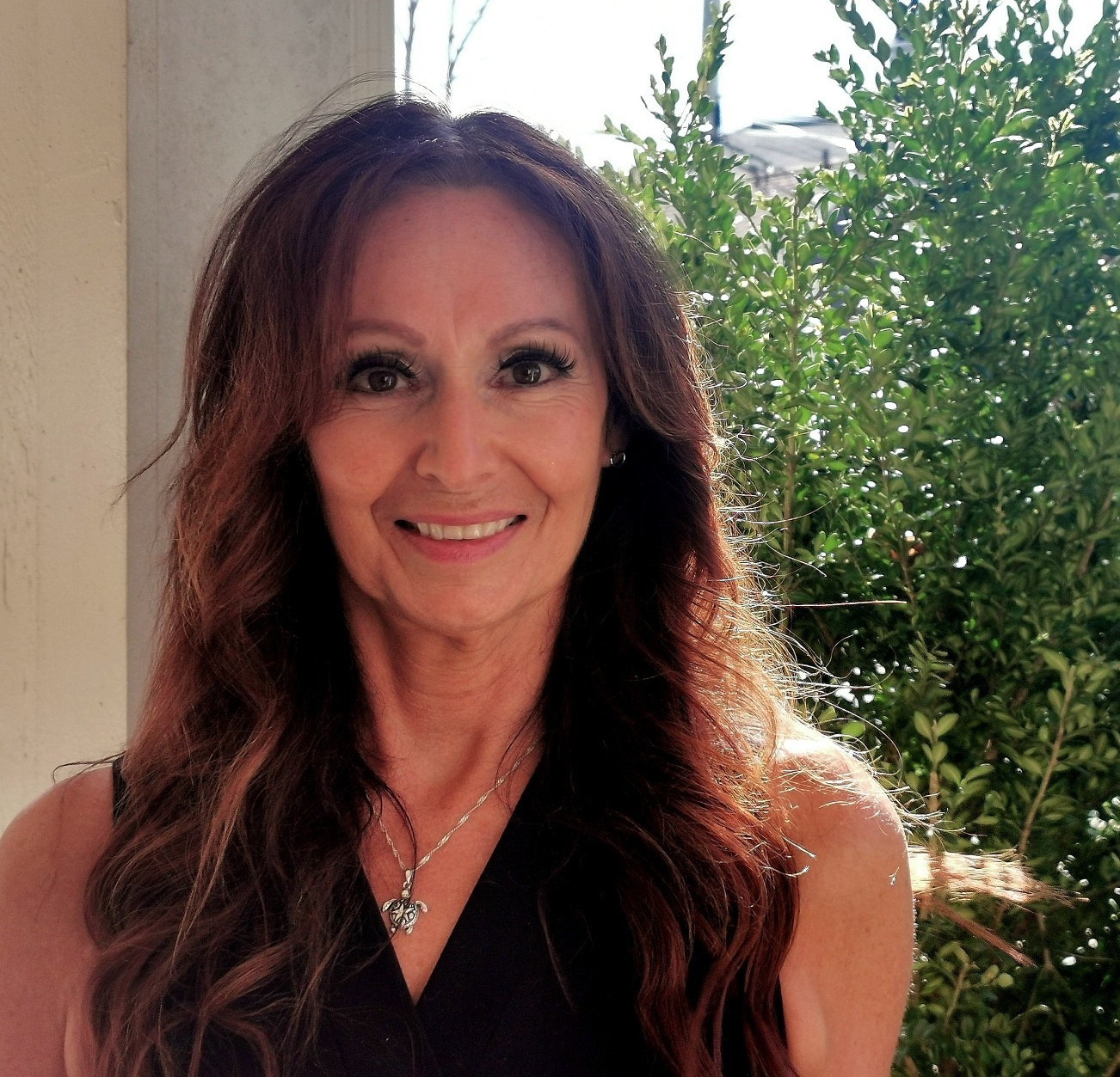 A woman with long brown hair and makeup, smiling outdoors near a house and green bushes.
