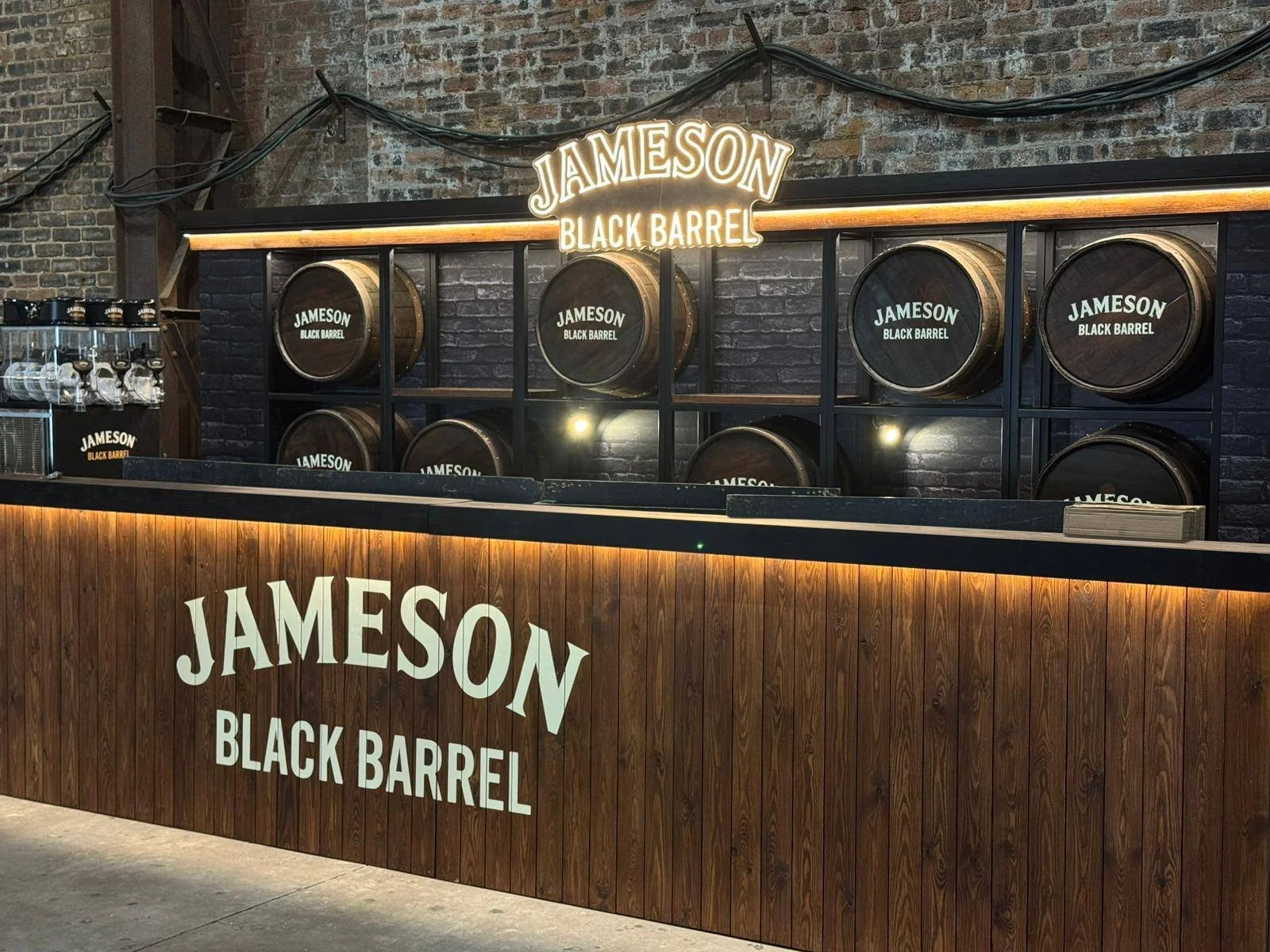 Bar with wooden front bearing the words "Jameson Black Barrel" in white, and barrels labeled "Jameson Black Barrel" arranged on a black rack behind the bar. There is a neon sign above the barrels that reads "Jameson Black Barrel." The background has 