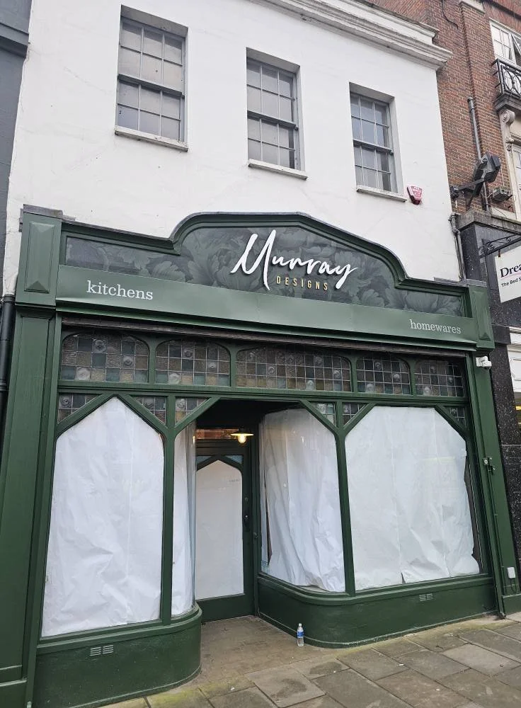 A storefront with green trim and large windows covered with white paper, with a sign that reads 'Murray Designs' and mentions 'kitchens' and 'homewares'.