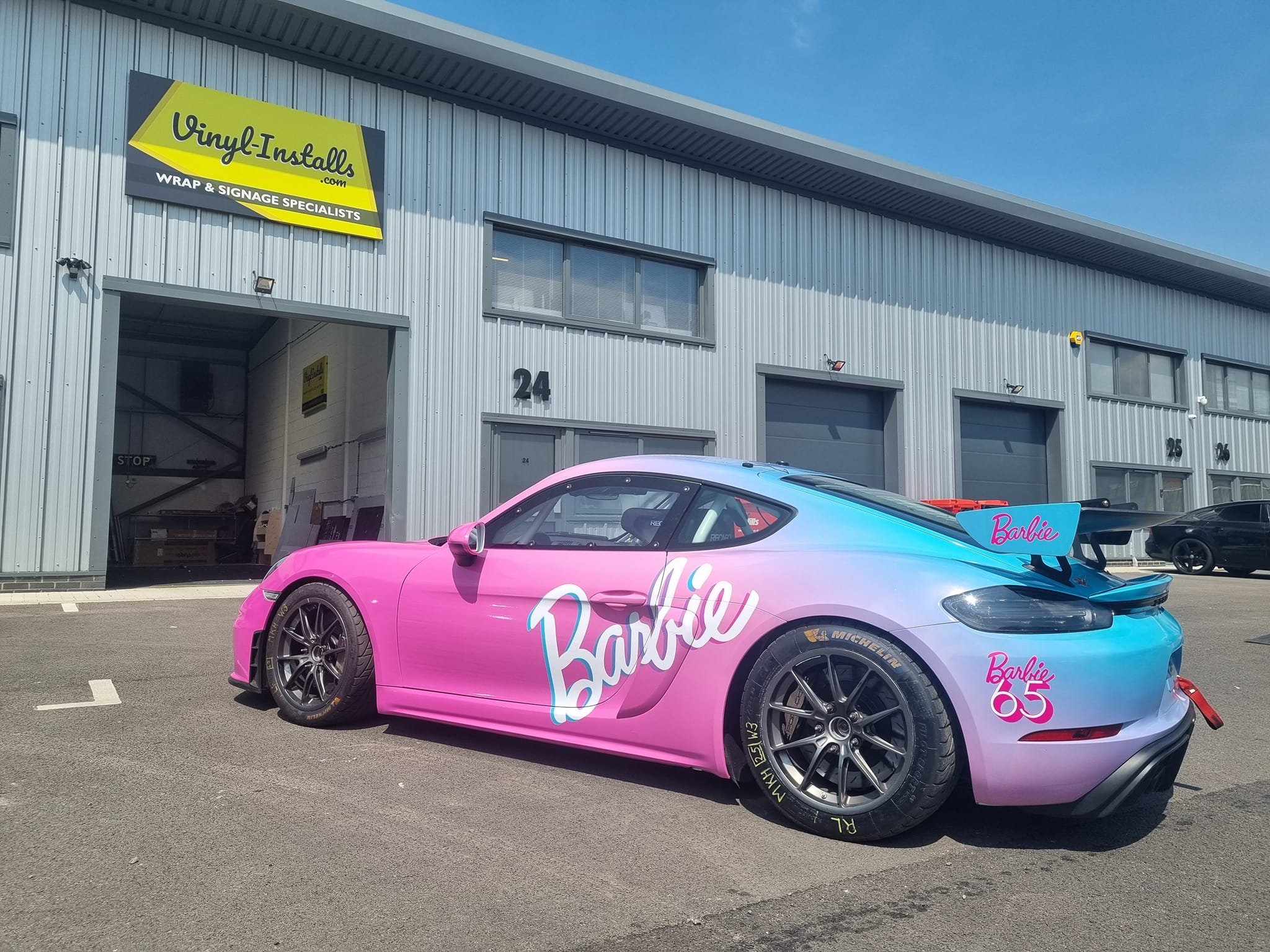 A sports car painted in pink and blue with Barbie branding and a large rear wing, parked outside a commercial building with a sign that reads 'Vinyl Install.com'.