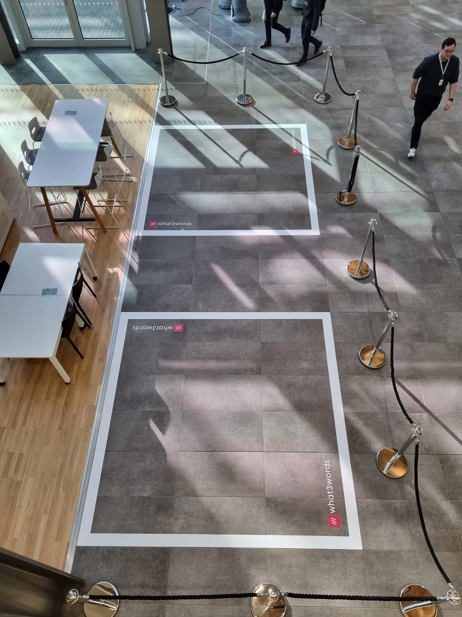 View of an indoor area with two empty interview or photo frames on the floor, marked with The What 3 Words locations, surrounded by black velvet ropes and stanchions. There are people walking in the background, and a corner with tables and chairs on 