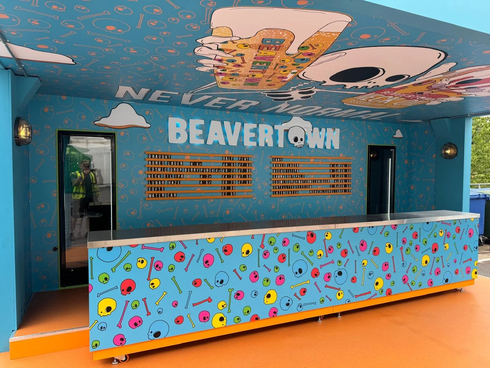 Colorful beverage stand with skeleton and skull motifs, menu listing drinks and prices, decorated with skulls and bones on the front, inside a building or event space.