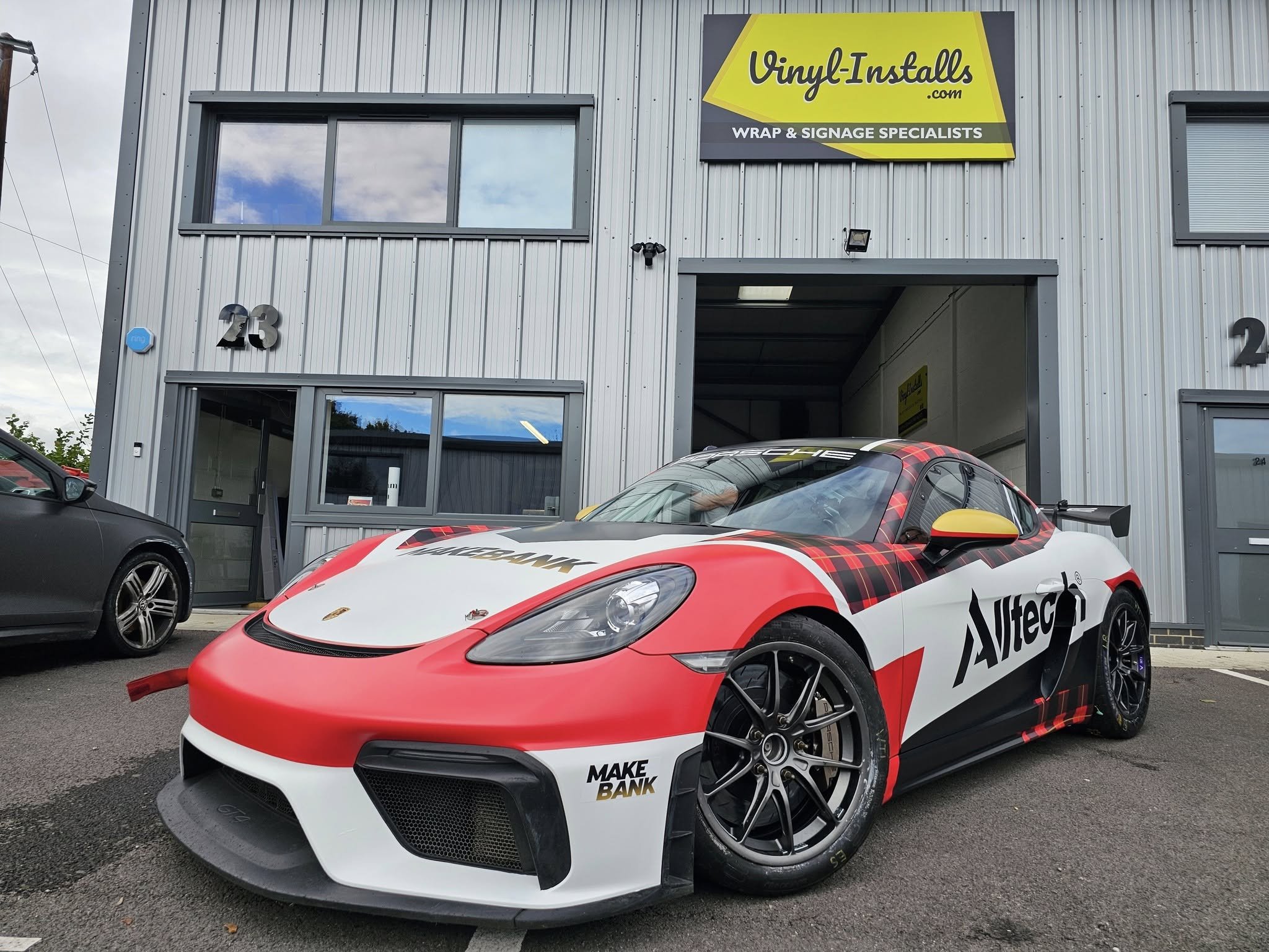 A sports car painted with a racing livery featuring sponsor logos parked outside a building with a sign for Vinyl-Installs.com, a wrap and signage specialist.