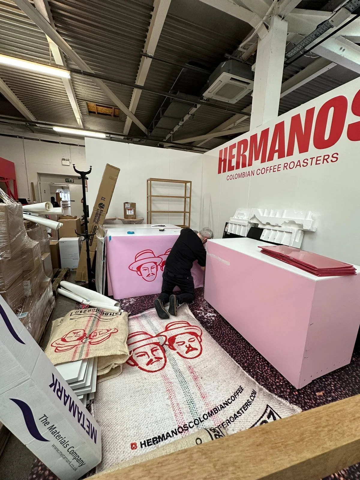A worker kneeling on the floor of a store or warehouse, arranging a pink display counter with Hermanos Colombian Coffee Roasters branding. There is a large white wall with red lettering that says HERMANOS and Colombian Coffee Roasters. Several stacke