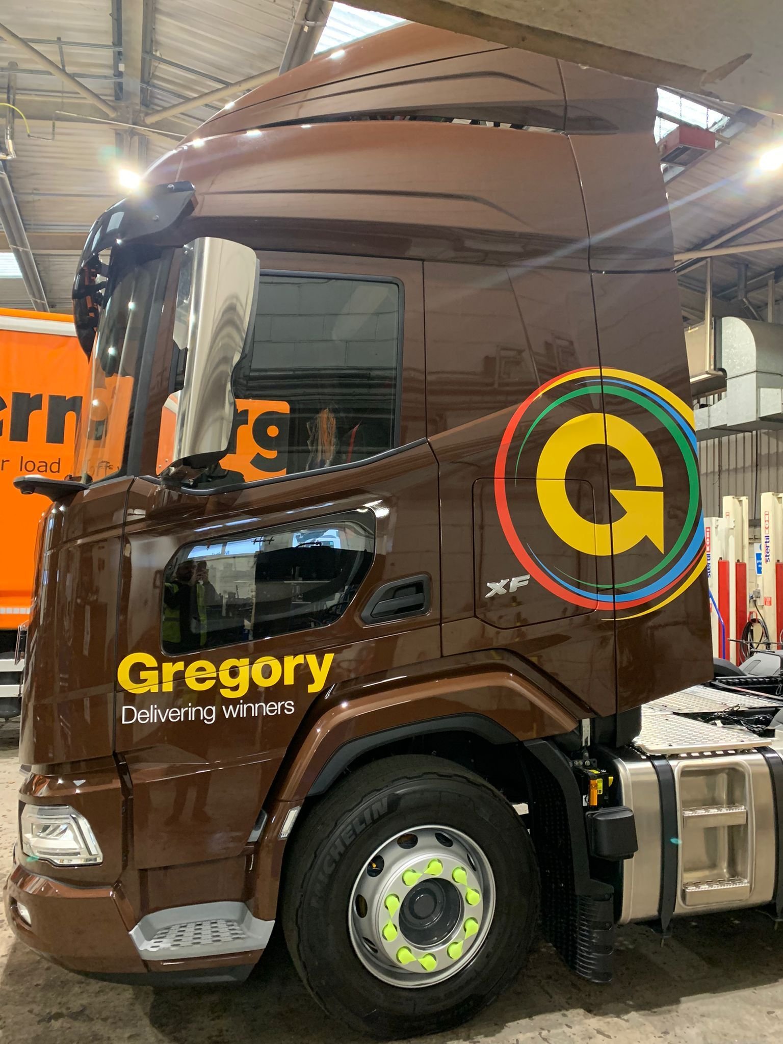 A brown semi-truck with the company name 'Gregory' and the slogan 'Delivering winners' painted on the side. The truck has a large circular logo with a yellow 'G' and colorful rings around it.