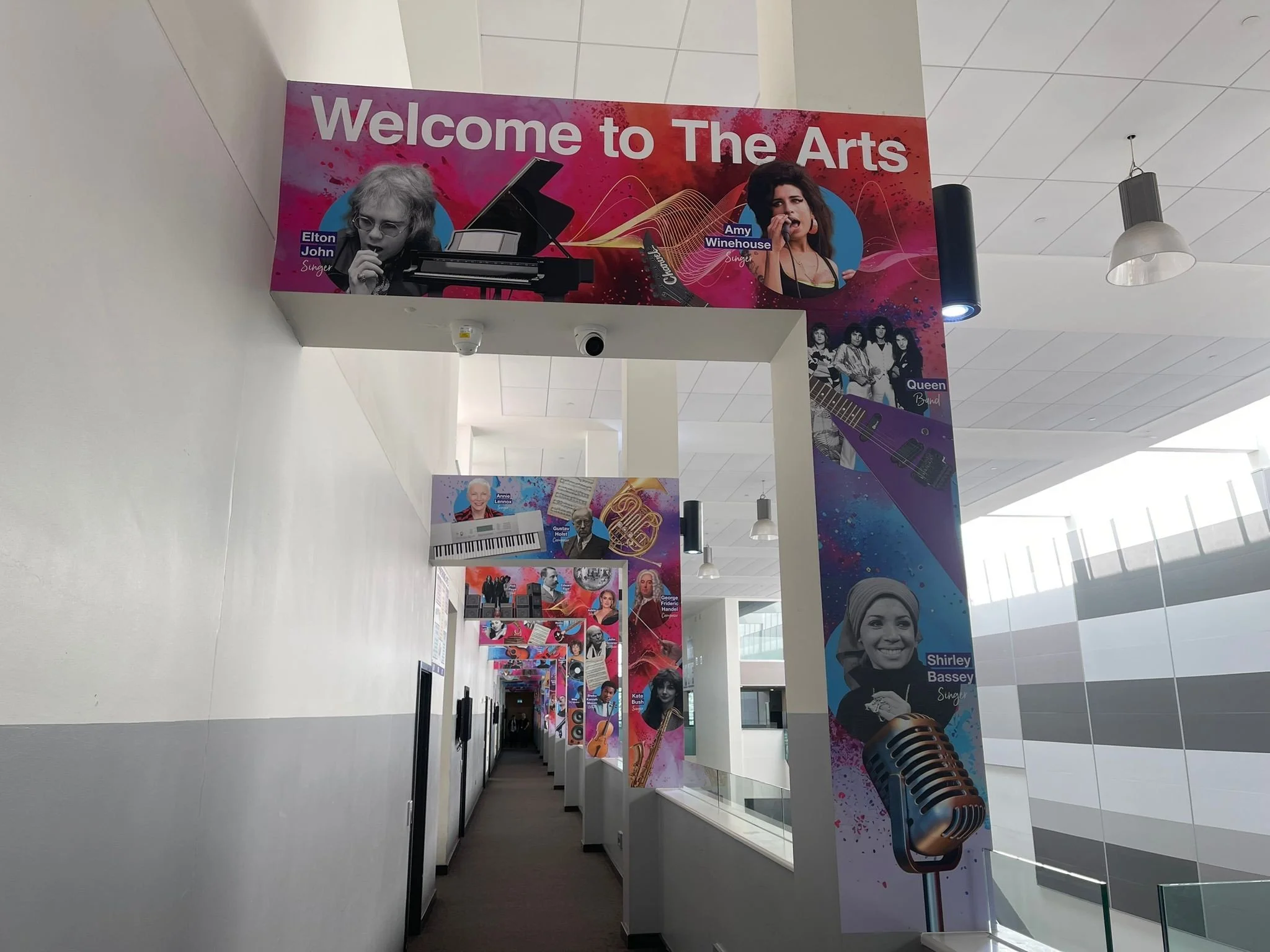 Colorful corridor with posters of musicians and singers including Elton John, Amy Winehouse, Queen, Shirley Bassey, and others, welcoming visitors to 'The Arts'.