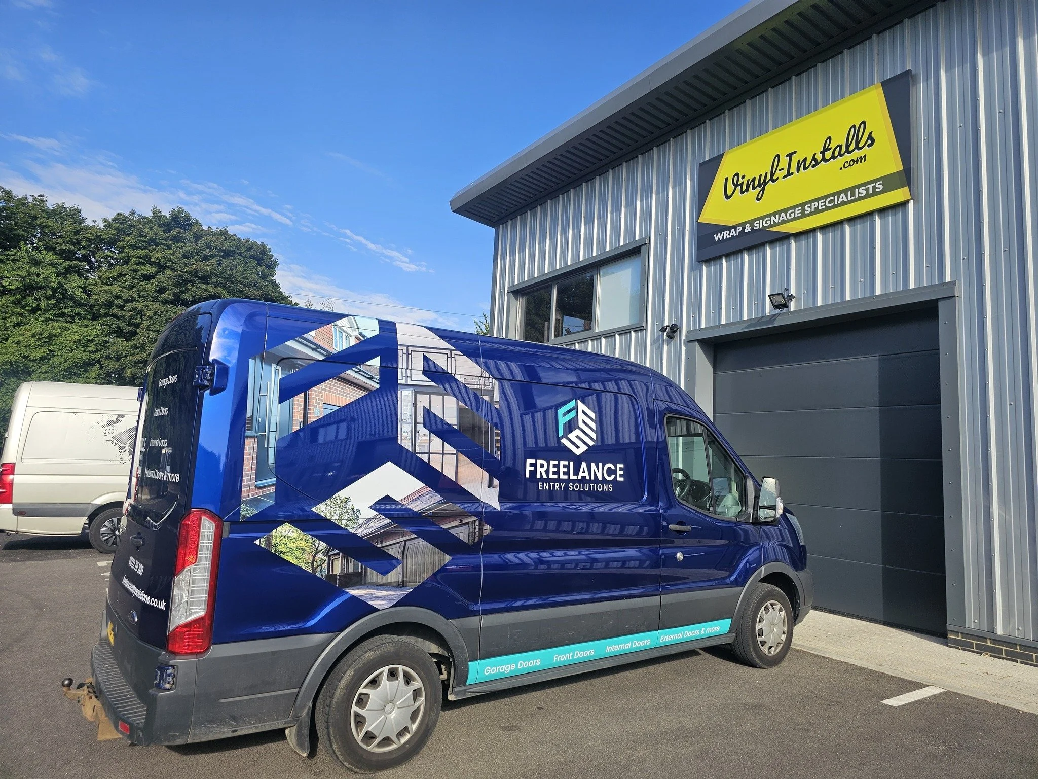 A blue van with branding for Freelance Entry Solutions, parked outside a commercial building with a yellow sign for Vinyl Installs, a wrap and signage specialist.