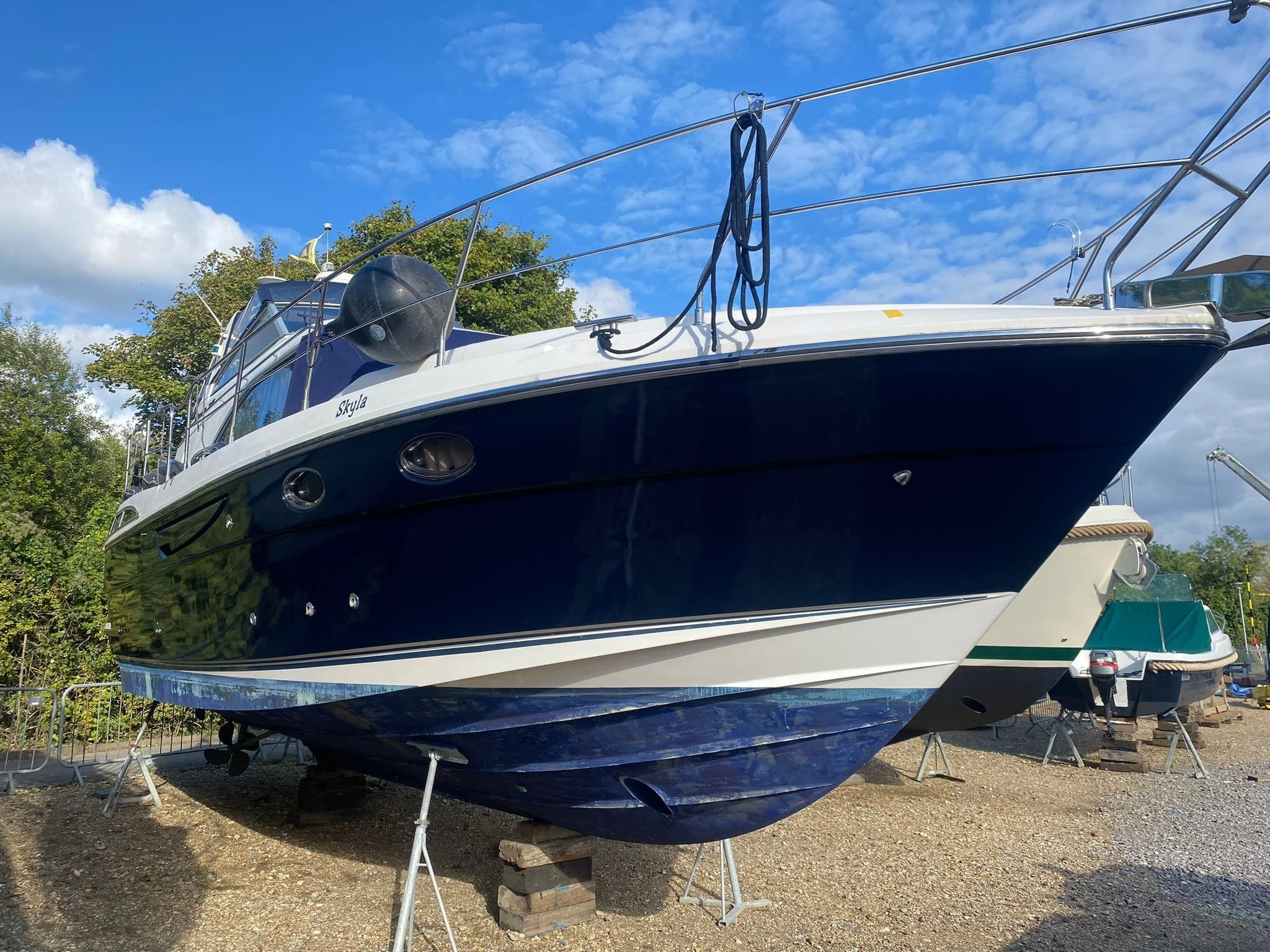 A dark blue and white yacht named Skyline on land, supported by wooden blocks and metal stands, with trees and a blue sky with clouds in the background.