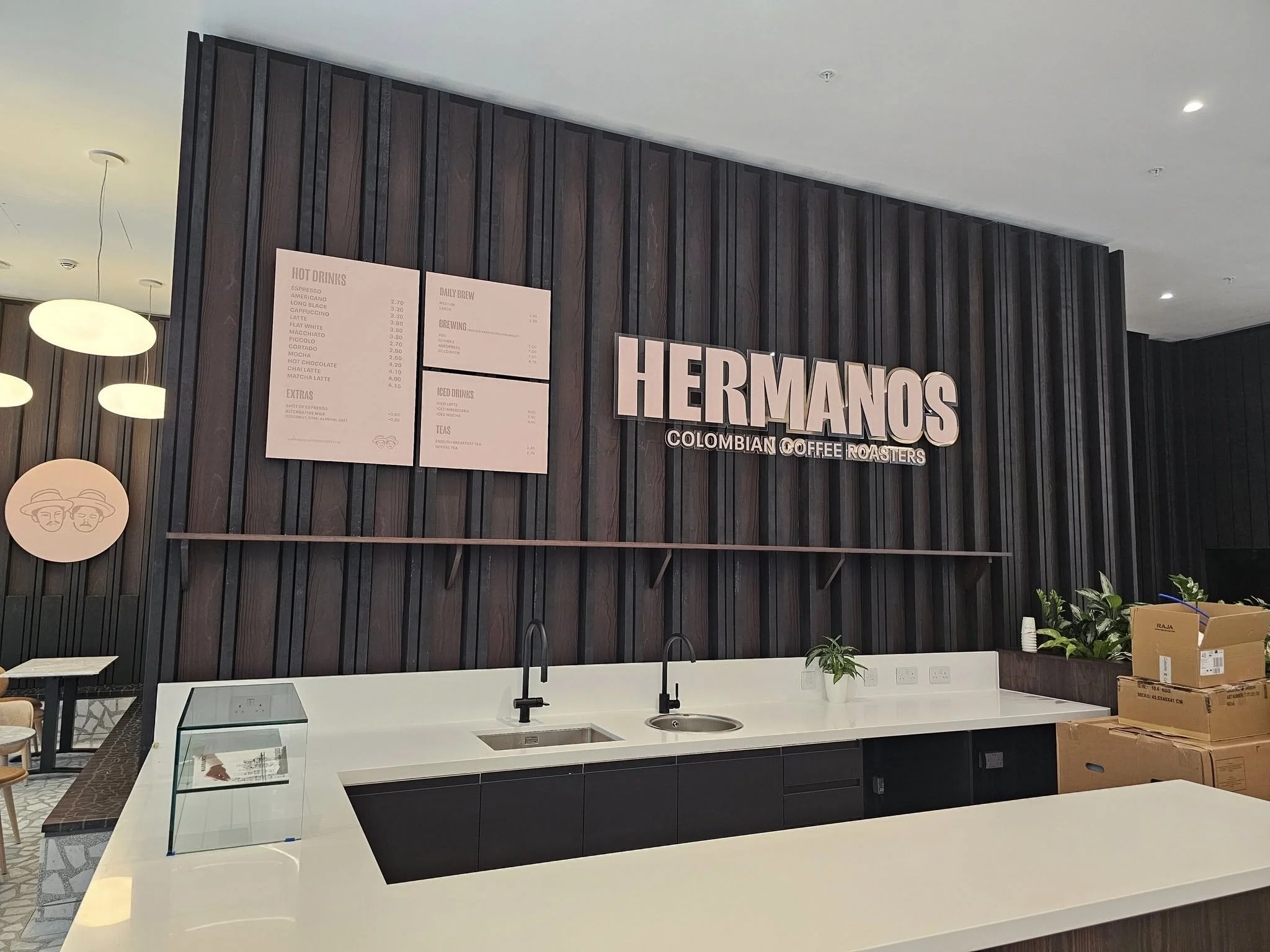 A person wearing a black cap with a stylized 'D' logo, sitting at a counter in a coffee shop, facing a menu board with a coffee cup illustration.