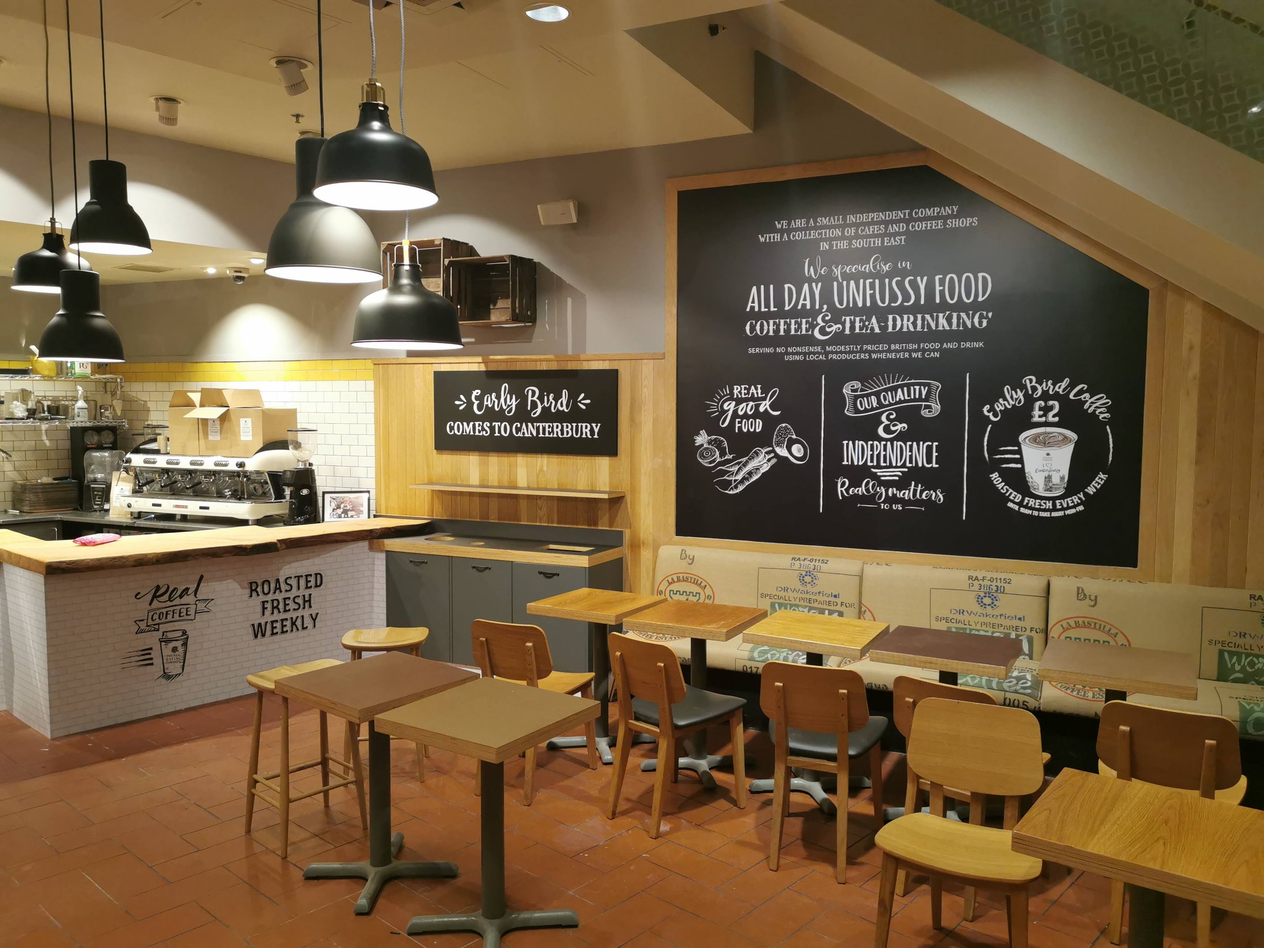 Inside a cozy coffee shop with wooden tables and chairs, black pendant lights, a large chalkboard menu with white handwritten text, and a counter with coffee machines and baked goods.