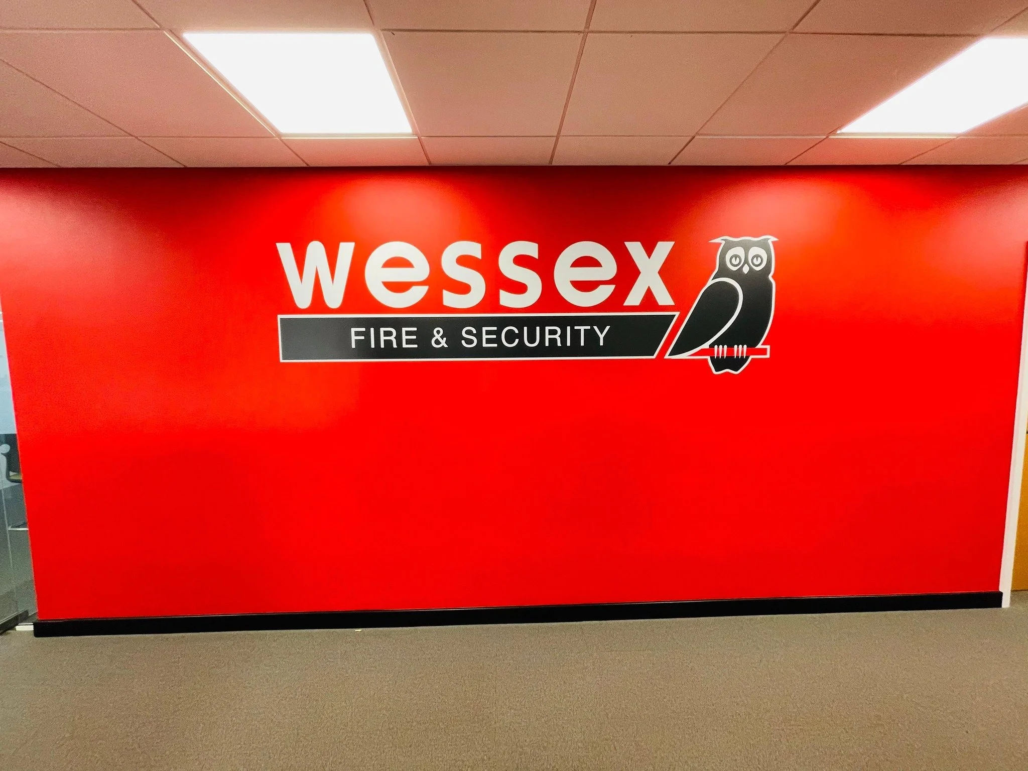 Red wall with large signage for Wessex Fire & Security, featuring a black and white owl holding a red and white stick.
