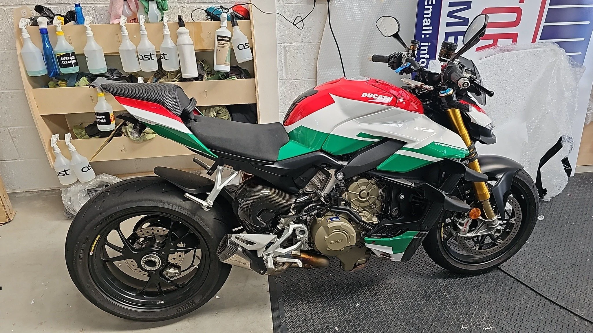 A Ducati Panigale V4 motorcycle in green, white, and red colors, with black wheels, gold front forks, and a carbon fiber exhaust, is parked indoors on a rubber mat.