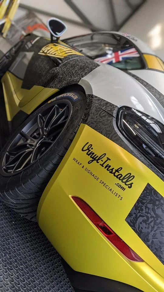 A yellow and black race car with a decal that reads Vinyl-Insalls.com, a wrap and signage specialist, parked indoors on a rubber mat with a small American flag sticker visible through the windshield.