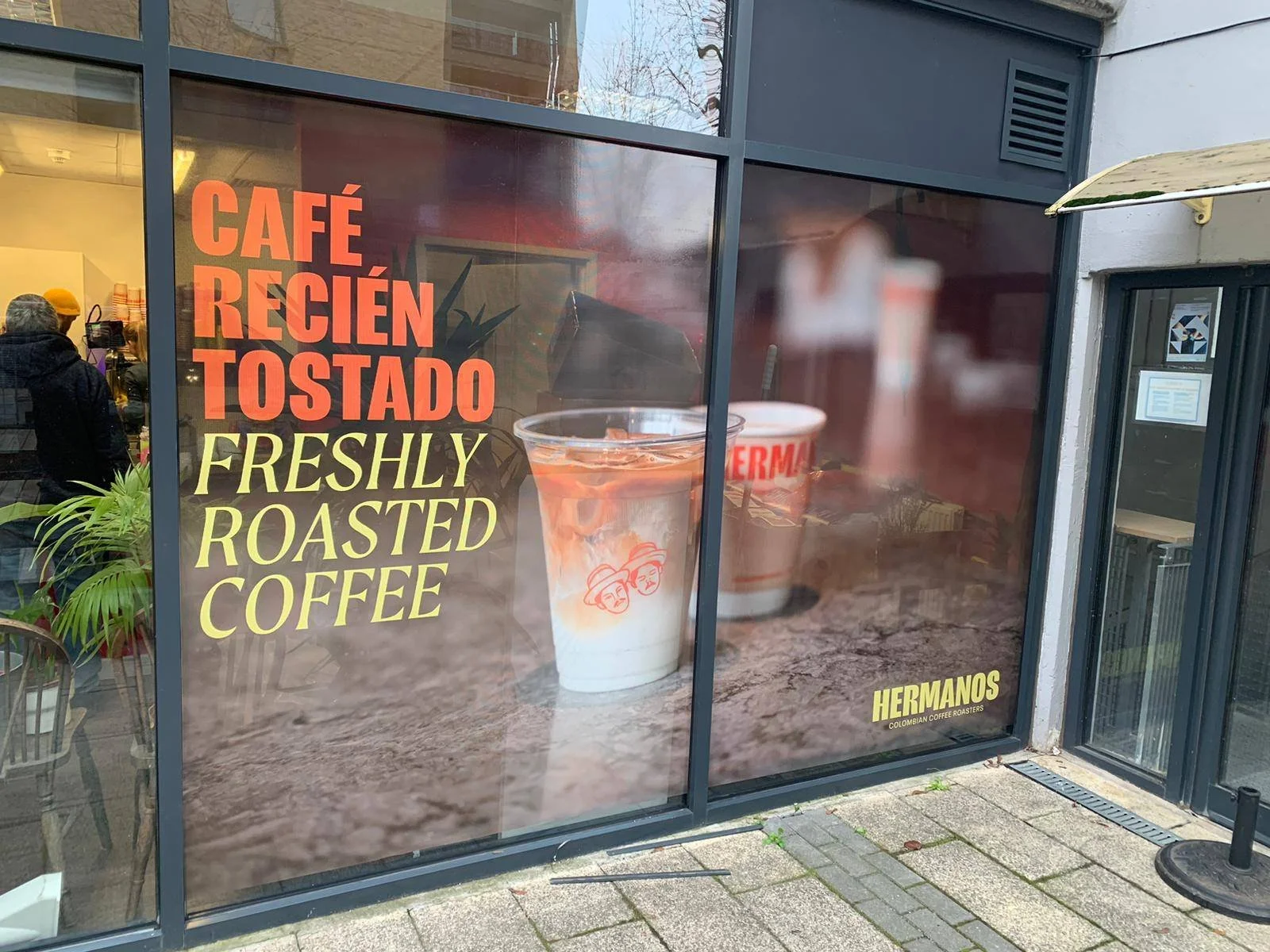Cafe window display with large poster advertising "Freshly Roasted Coffee" in English and Spanish, featuring an image of a glass of coffee with milk and a cup behind it. The cafe name is Hermano's Colombian Coffee Roasters.