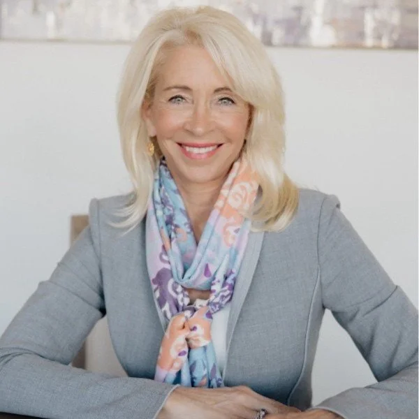 A smiling middle-aged woman with blonde hair, wearing a gray blazer and a multicolored scarf, sitting at a desk in an office with large windows.