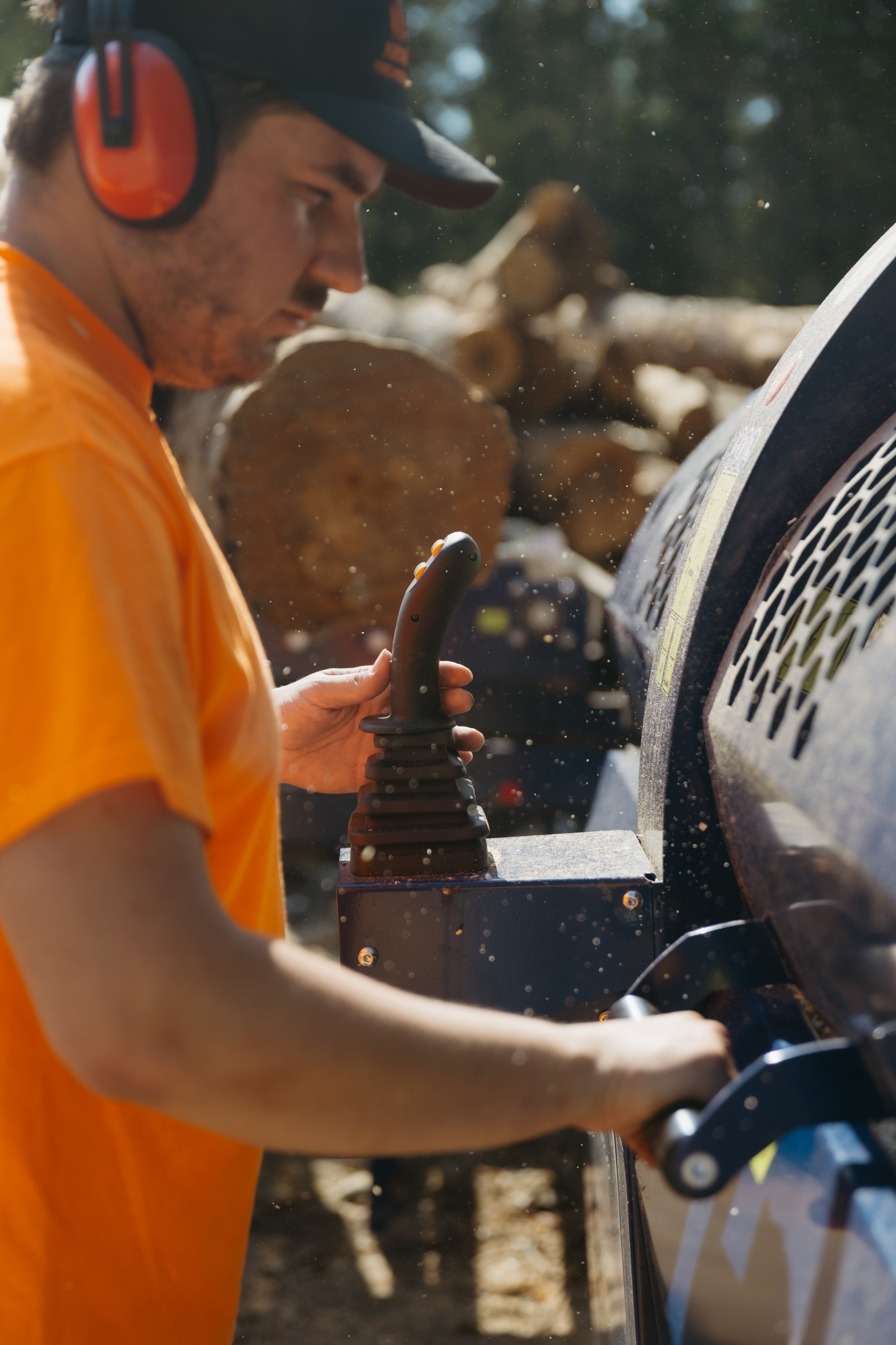 A young man with light skin and short dark hair, wearing an orange shirt, orange and black headphones, and a dark baseball cap, is operating a piece of heavy equipment with his right hand, while working outdoors near cut logs with a blurry background of trees.