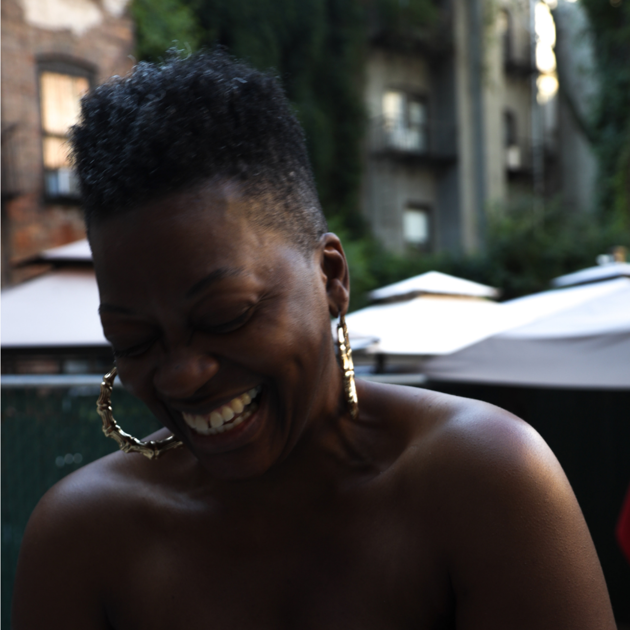 A woman with short curly hair and large gold hoop earrings dancing and smiling outdoors, with buildings and umbrellas in the background.