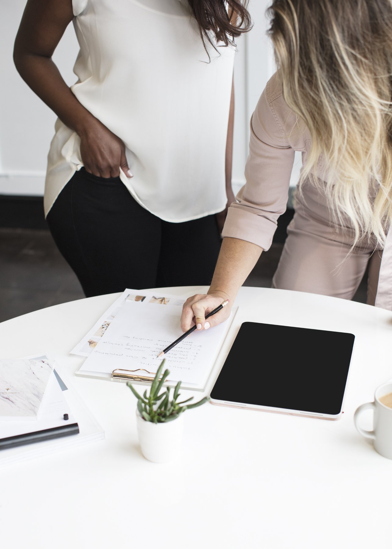 Two women working at a white table with papers, a tablet, and a cup.
