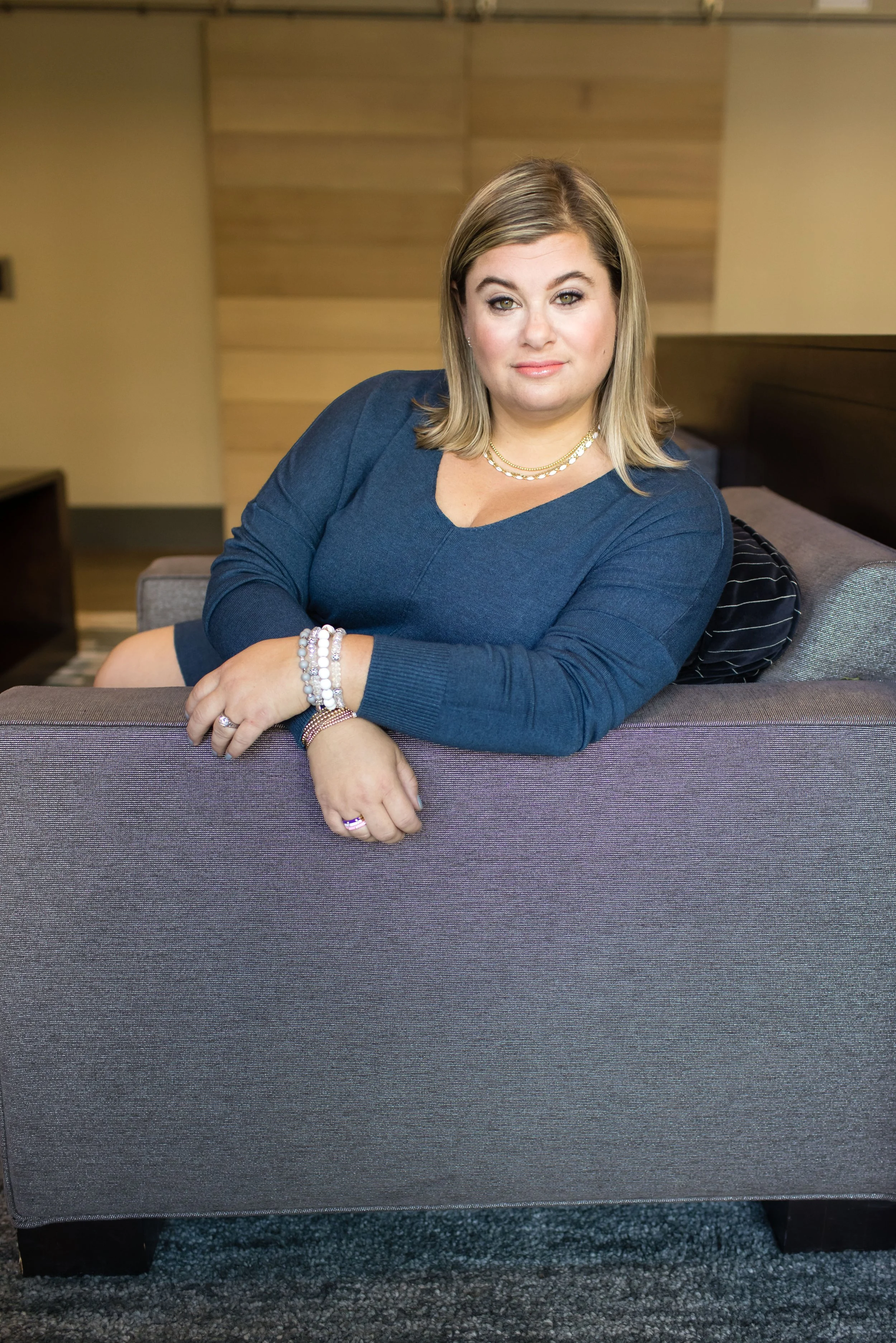 A woman with shoulder-length blonde hair, wearing a dark blue long-sleeve top and jewelry, is sitting on a gray sofa with a relaxed pose, in an indoor setting with a wood-paneled wall in the background.