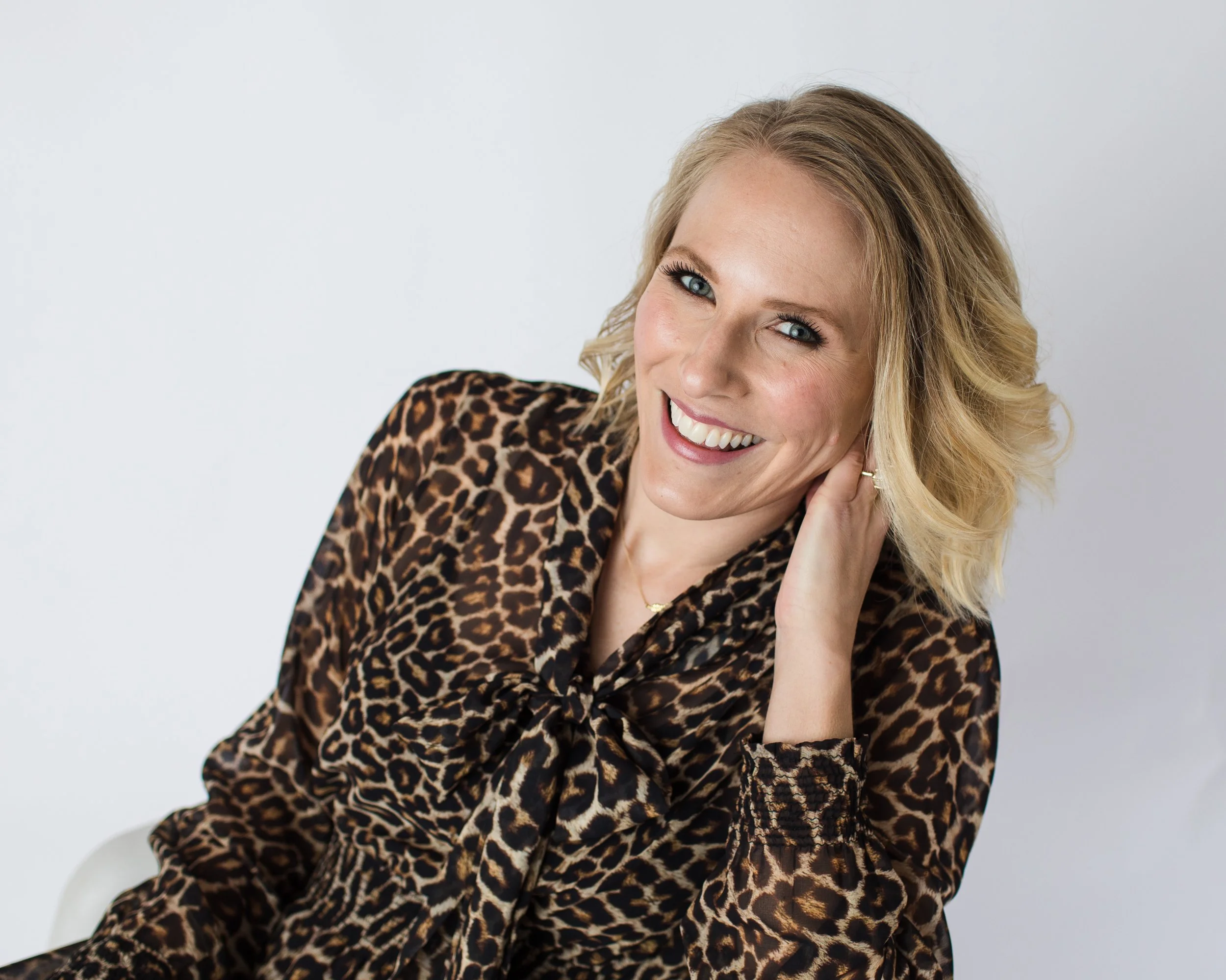 A blonde woman smiling and touching her ear, wearing a leopard print blouse against a white background.