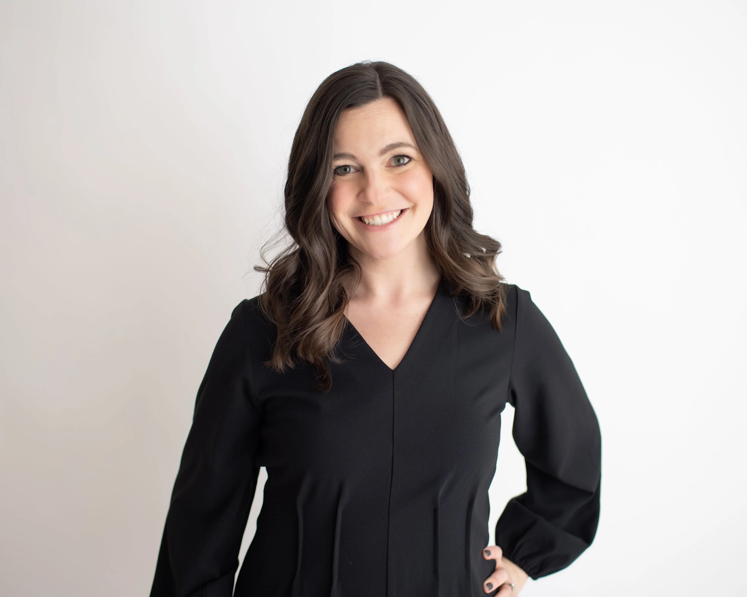 A woman with wavy brown hair, wearing a black dress, smiling with her hand on her hip, standing against a plain white background.