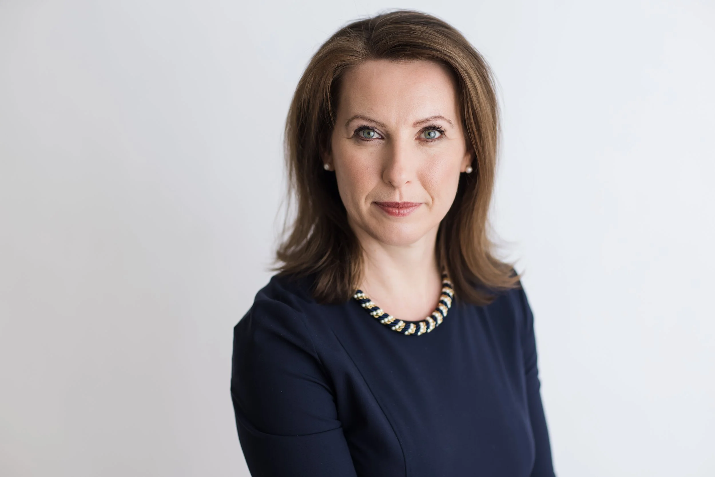 Professional woman with shoulder-length brown hair, blue eyes, wearing a navy blue dress and a beaded necklace, standing against a plain white background.