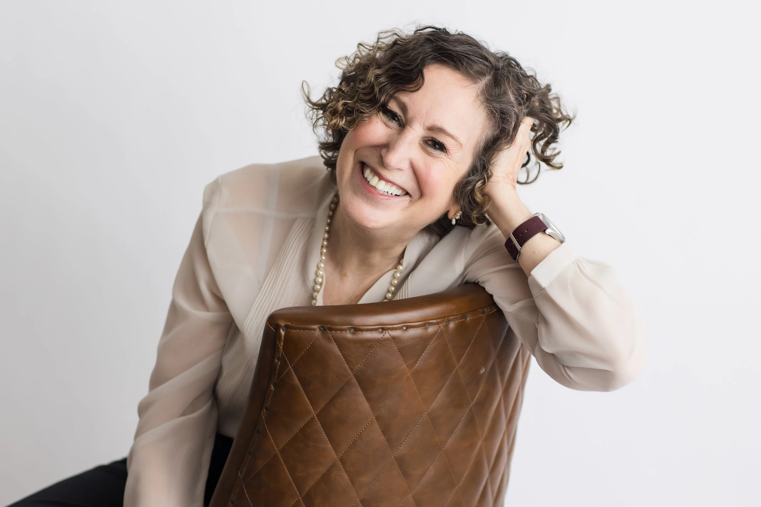 A woman with curly hair smiling and leaning on a brown leather chair against a plain white background.