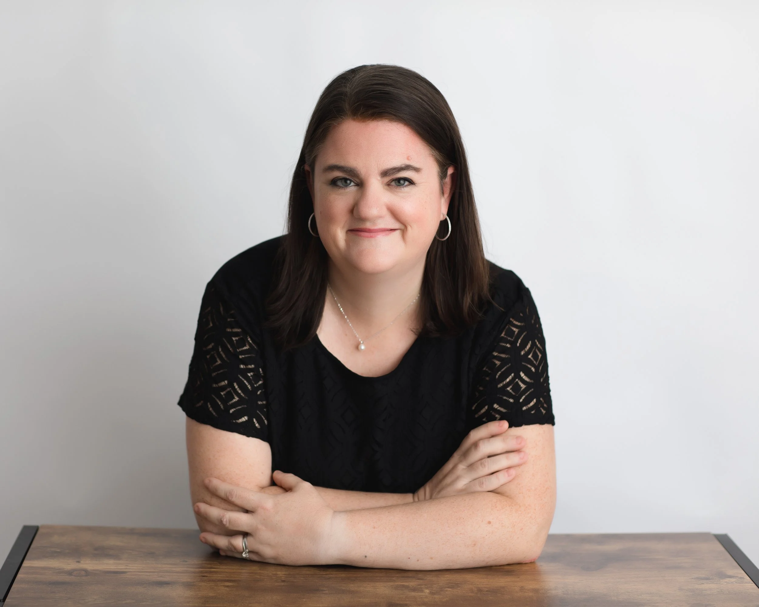 A woman with dark brown hair, wearing a black lace dress, sitting at a wooden table against a white background, smiling with her arms crossed on the table.